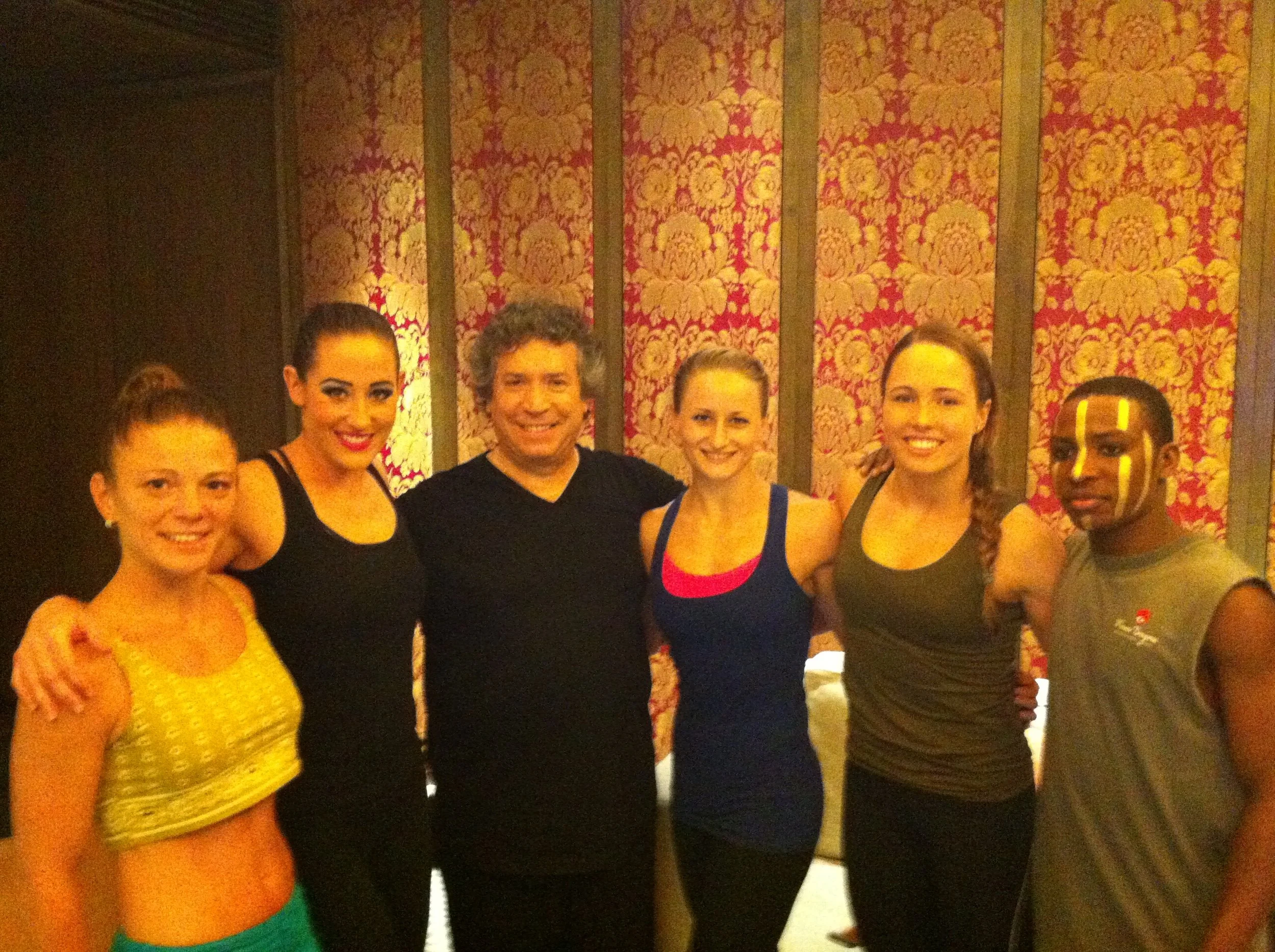 Group of five performers posing together with Franco Dragone indoors, with patterned red and gold wallpaper in the background. 