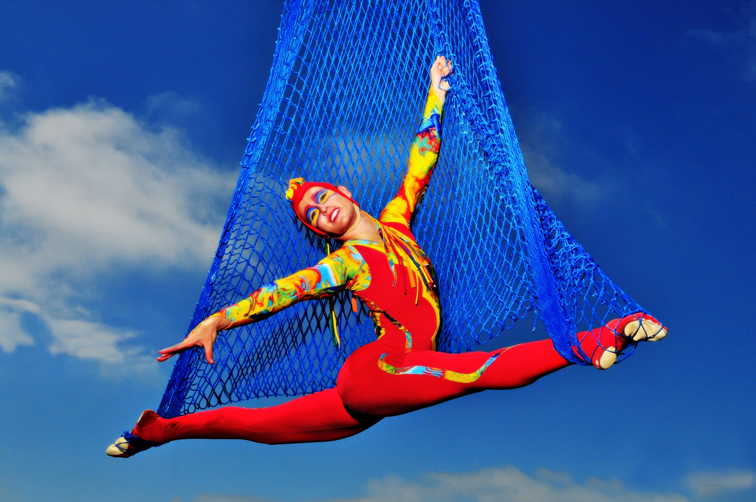 Circus performer in colorful costume performing a split inside a blue aerial net outdoors against a blue sky with clouds.