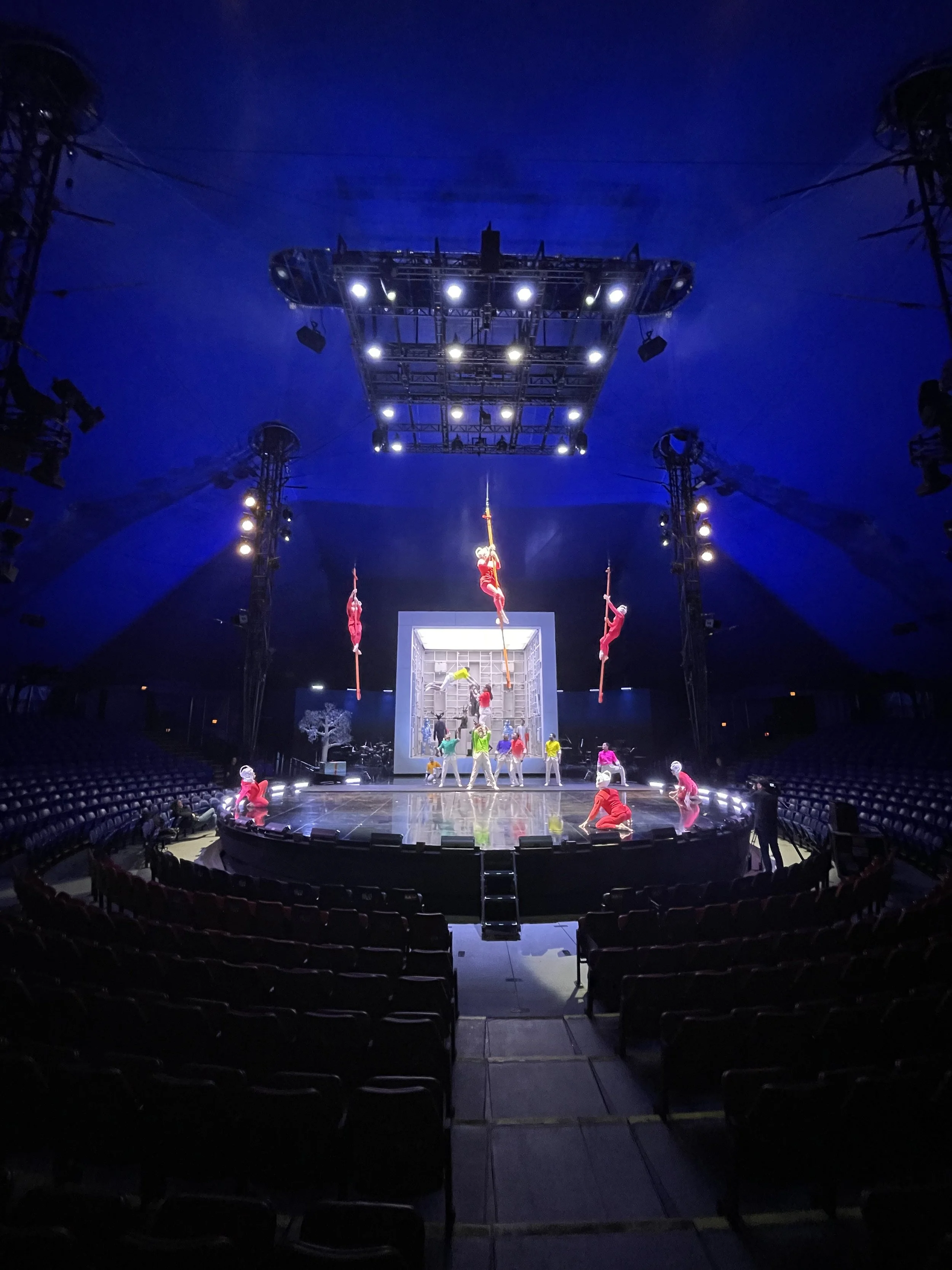 A circus ring with performers hanging from aerial poles and others kneeling on the floor, under bright stage lighting inside a large big top tent.