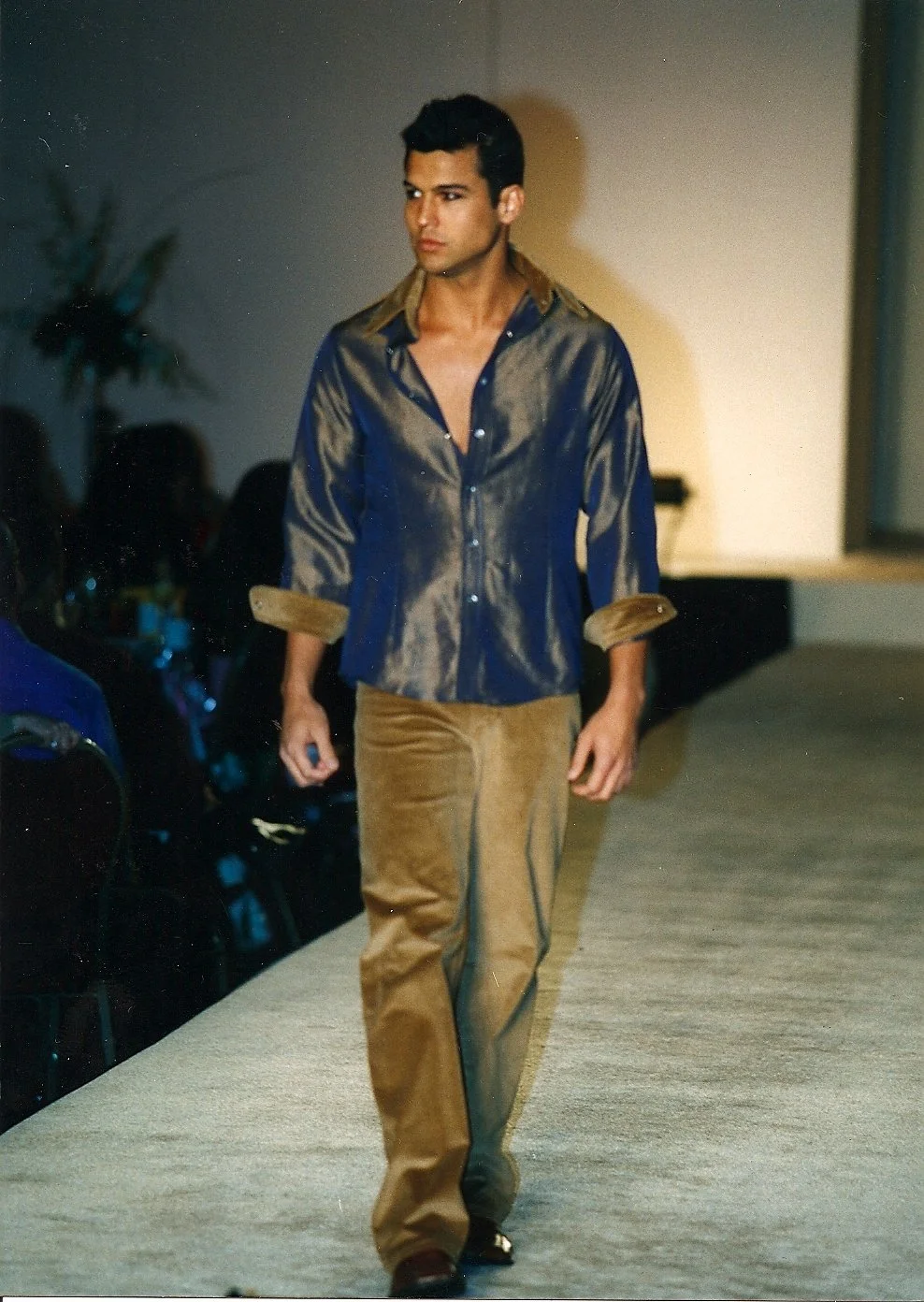 Male model walking on a runway during a fashion show, wearing a shiny purple long-sleeve shirt and brown pants.