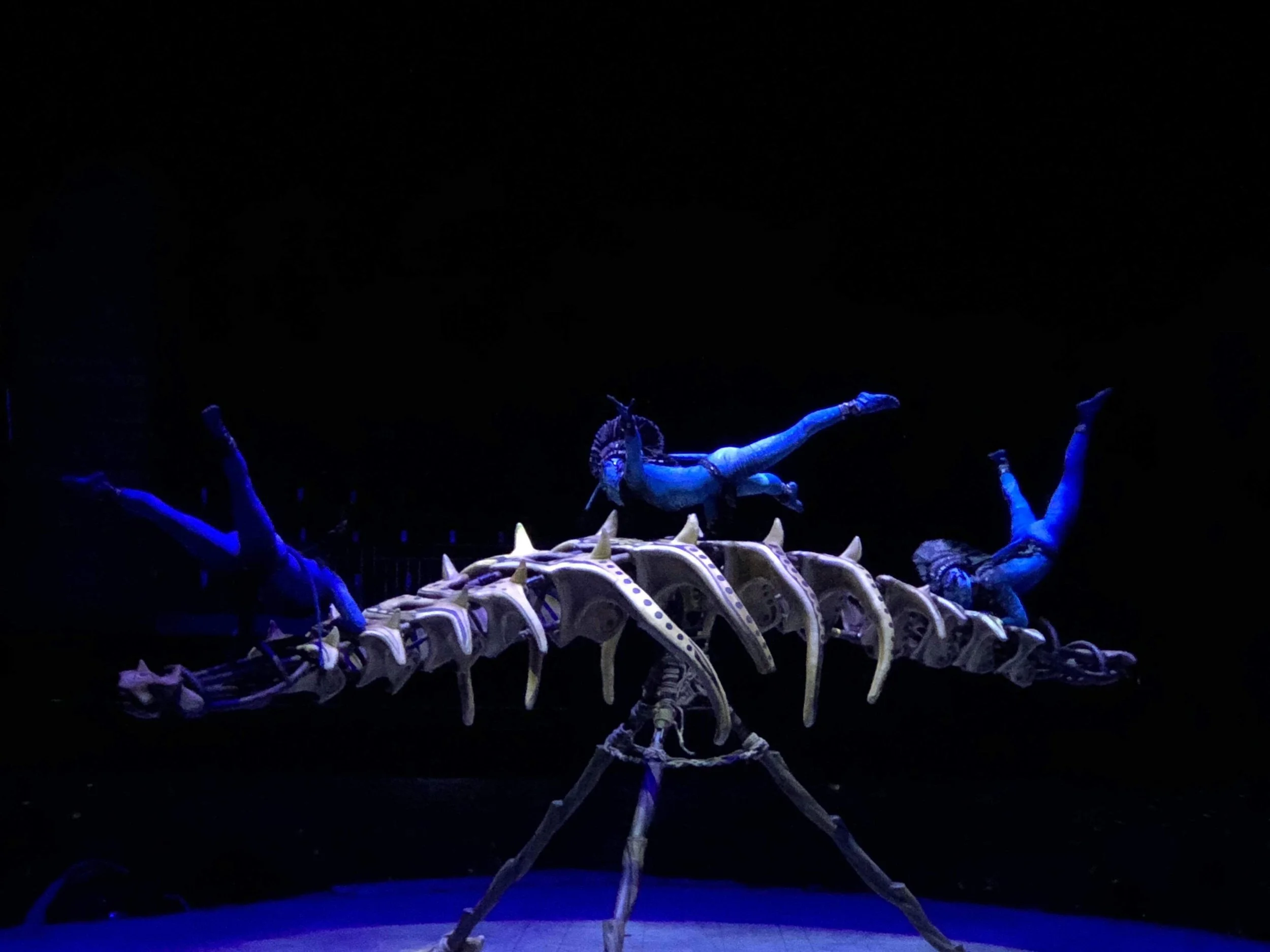 A performance of acrobats with Cirque du Soleil's Toruk wearing blue costumes in a dark setting with a mechanized skeleton acrobatic apparatus.