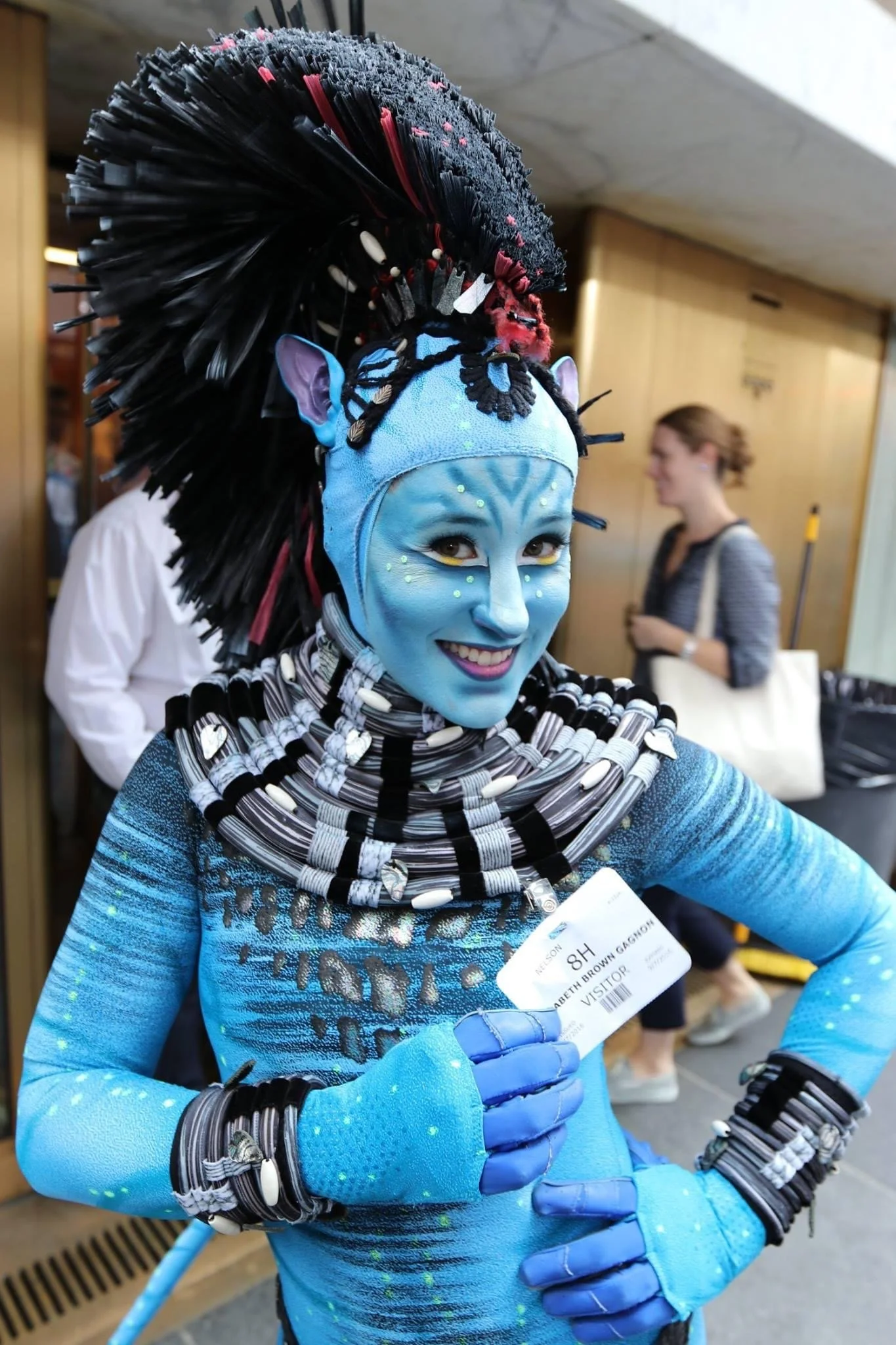 Woman dressed as Avatar character, painted blue with face markings, wearing elaborate headdress with black and red feathers, and body suit with painted details, holding a visitor badge.