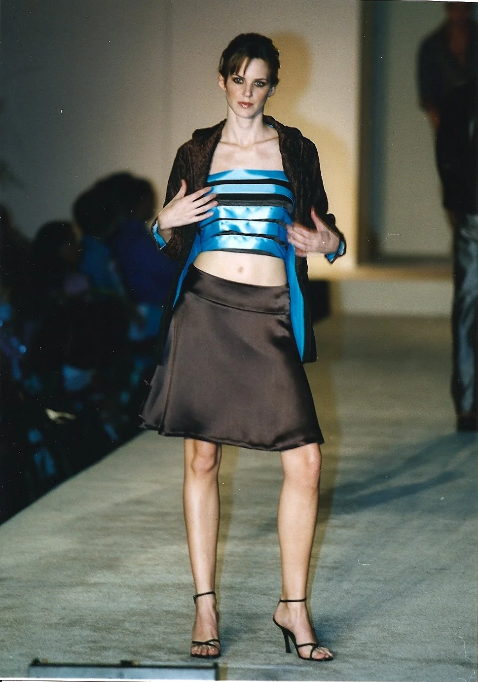 Woman walking on runway in a blue satin crop top, brown, textured jacket, brown satin skirt, and high heels at a fashion show.