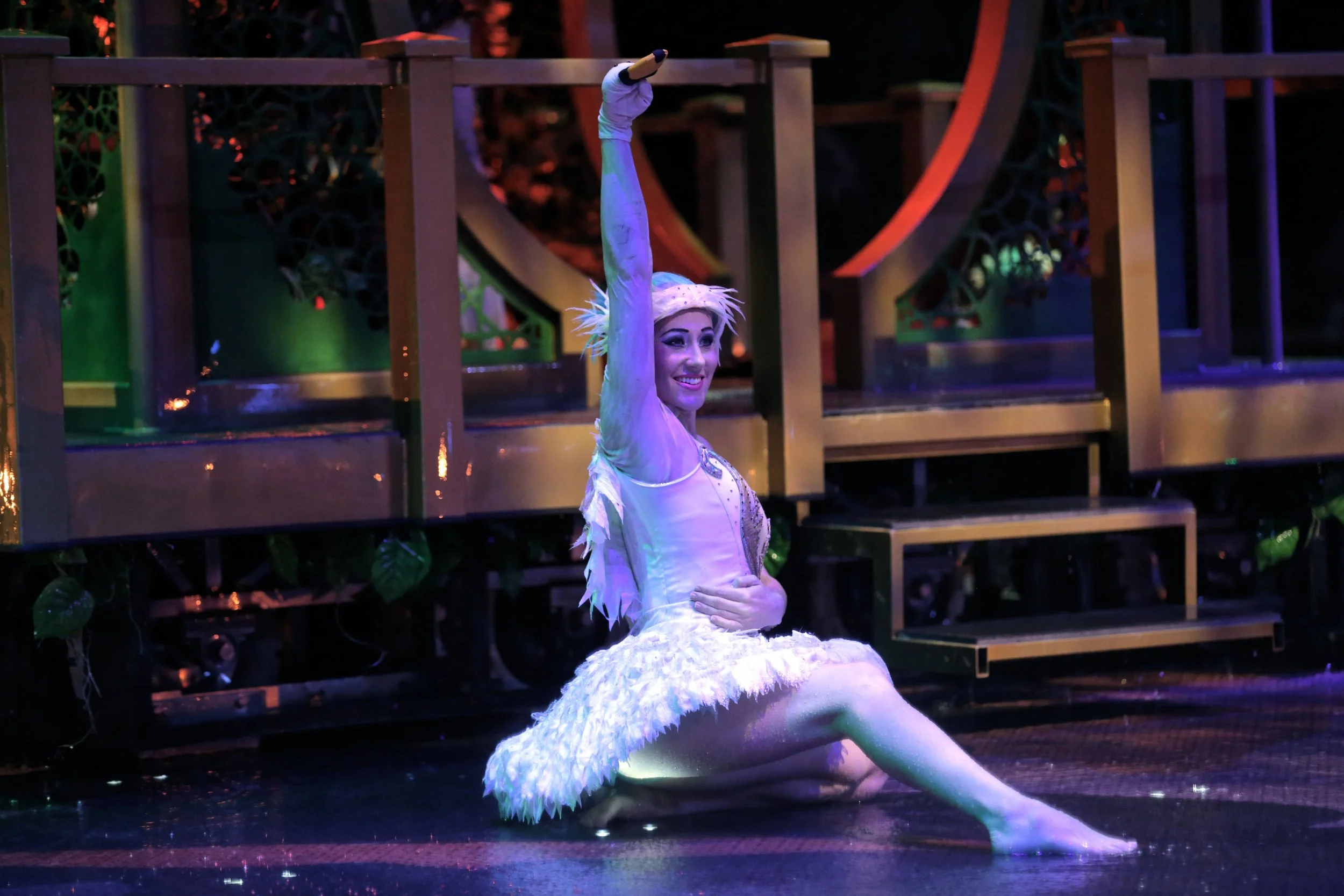 A female performer dressed as a bird or fairy, kneeling on stage with one arm raised and smiling, in a colorful theatrical setting.