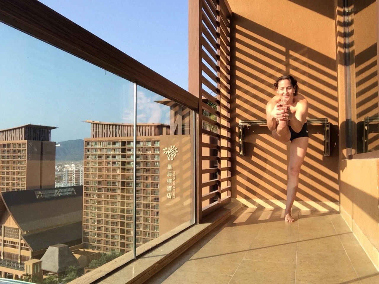 A woman in a black swimsuit stretching on a balcony overlooking city buildings with mountain in the background, sunlight casting striped shadows on the wall.