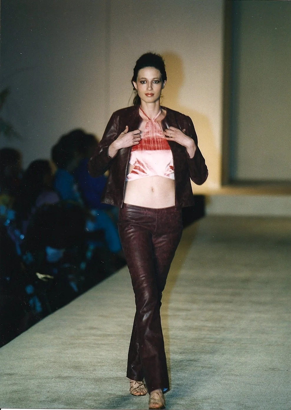 A female model walking down a runway during a fashion show, wearing a shiny, pink and burgundy outfit with a leather jacket and flared pants, holding the collar of her jacket with both hands.