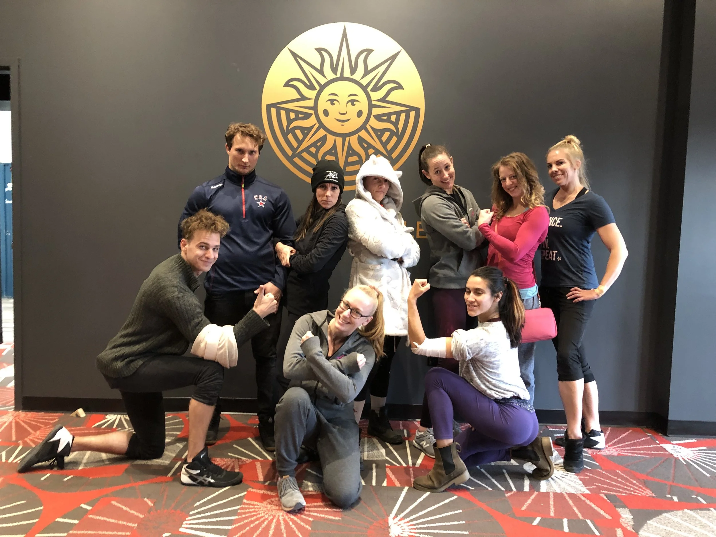 Group of nine women and men posing in front of a dark gray wall with a yellow sun logo, some flexing and smiling, in an indoor setting with a patterned carpet.