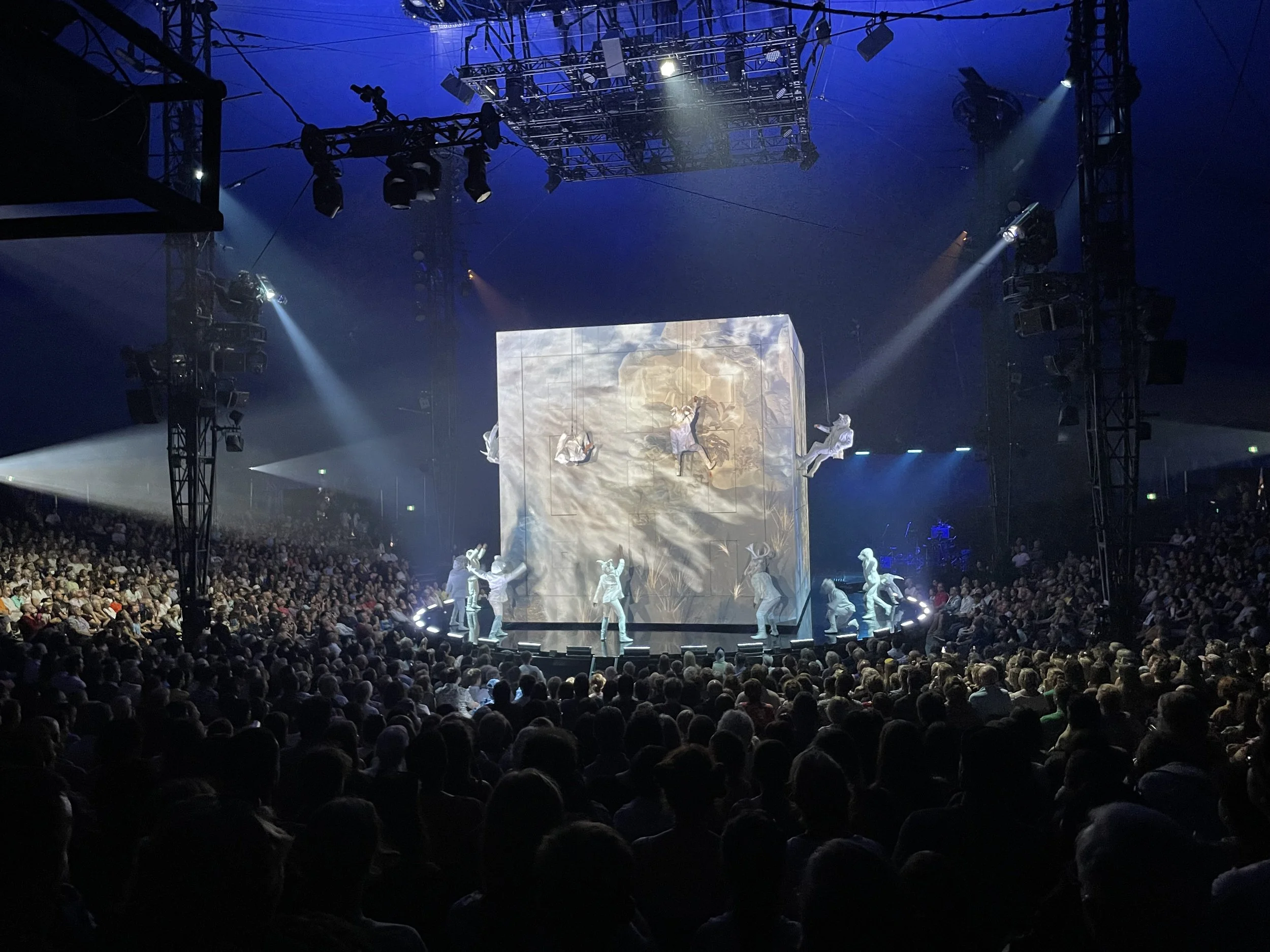 A circus performance inside a large tent with a stage, featuring a large set piece in the shape of a cube with a projected digital backdrop and performers in white costumes, with a large audience seated in front.