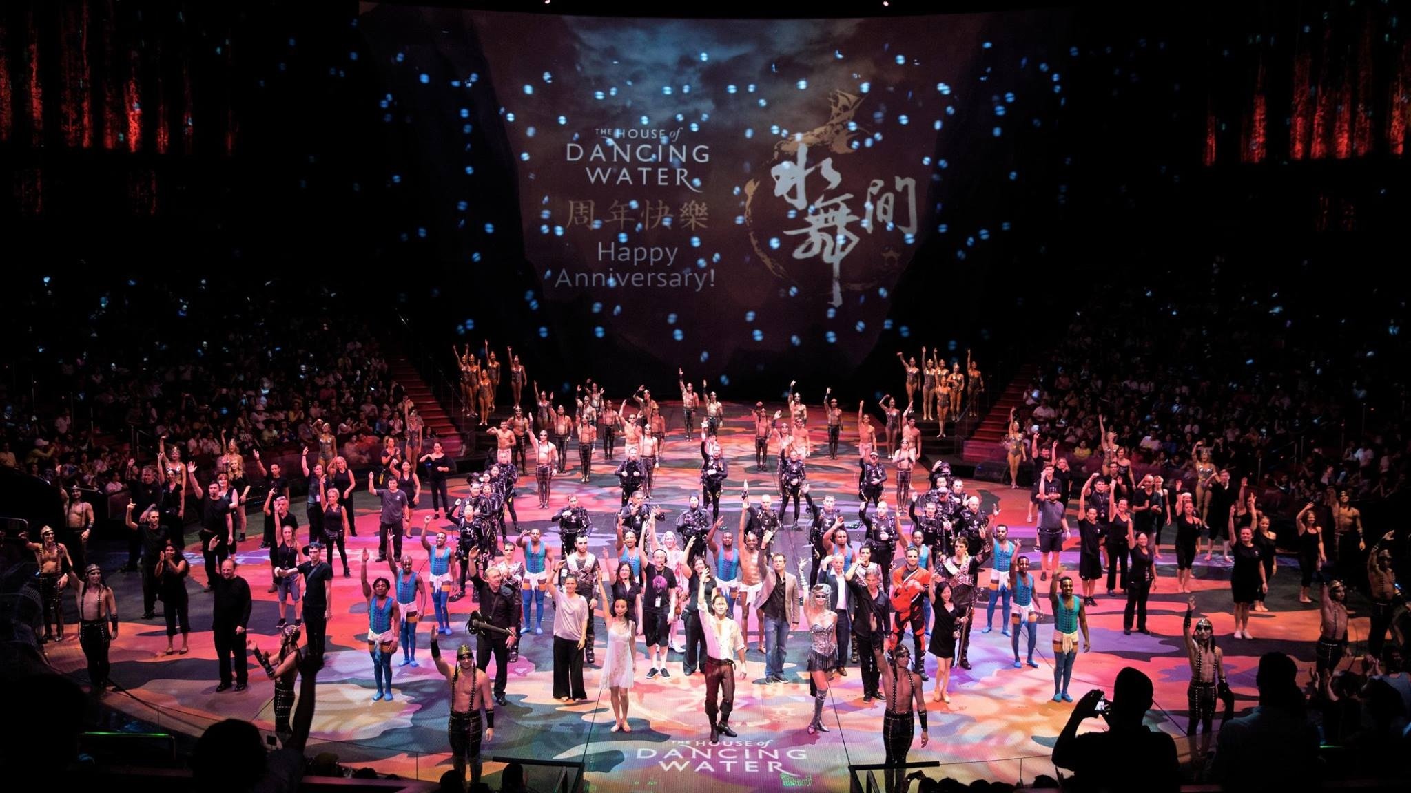 Large stage with many performers and dancers, audience seated around, colorful lighting, and a projection screen with text 'The House of Dancing Water' and 'Happy Anniversary!'