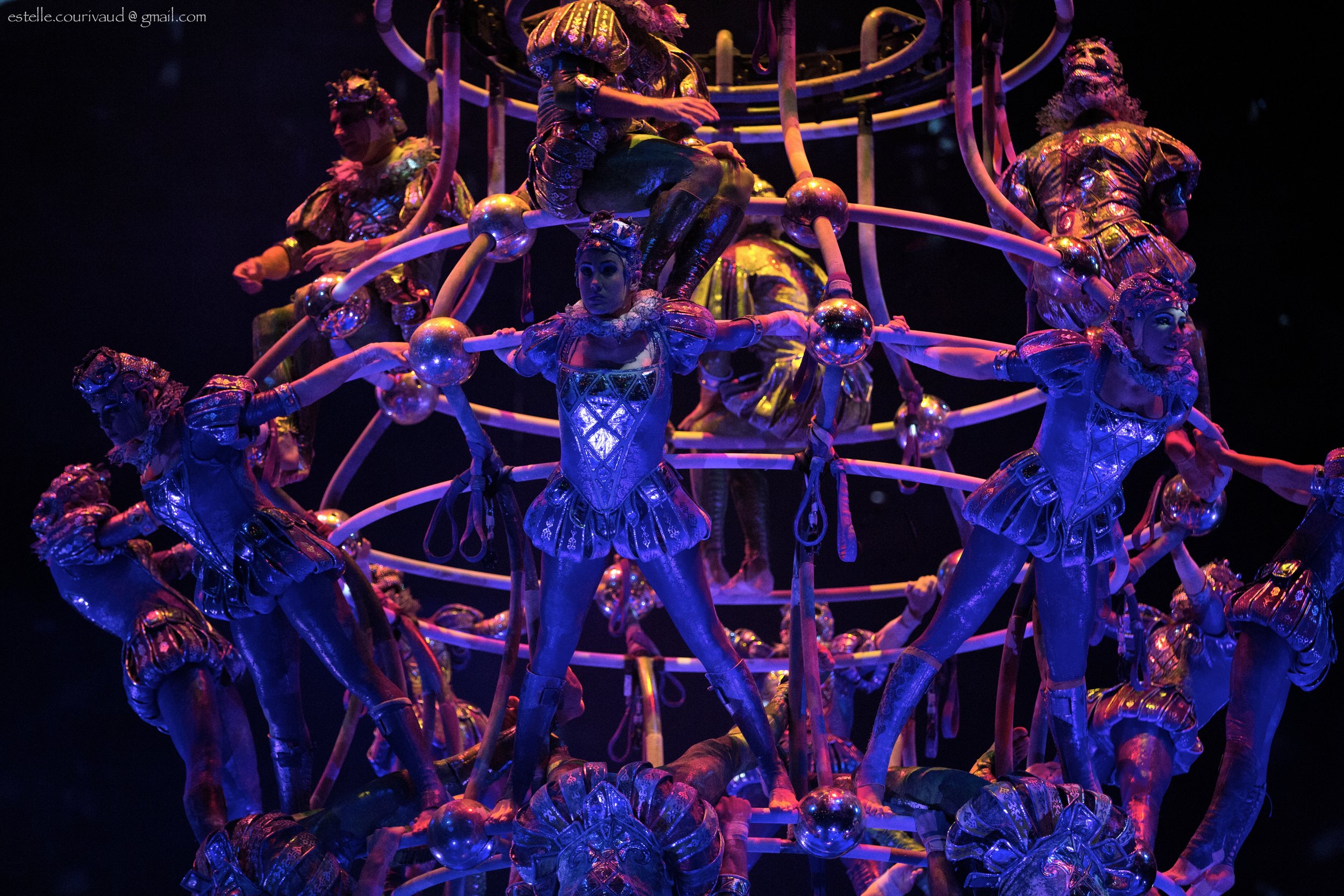 A group of performers dressed in elaborate, shiny costumes with metallic and reflective surfaces, performing on a multi-tiered, spherical structure with dynamic lighting that creates blue and purple hues.