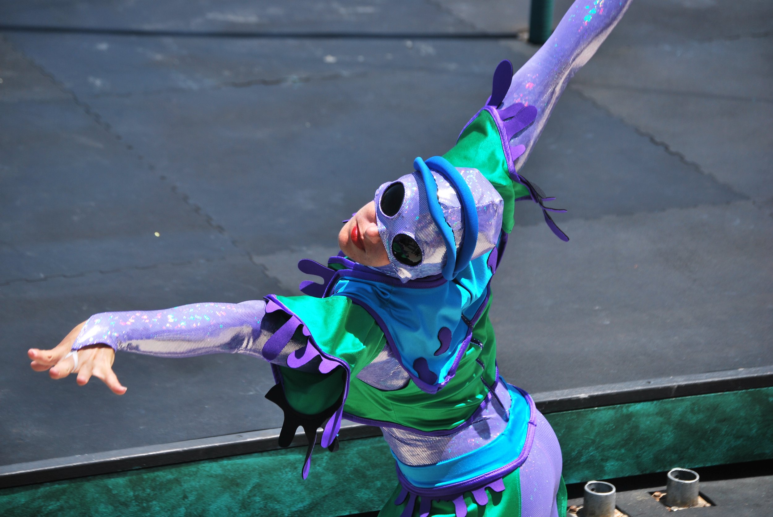 A female acrobat dressed in a colorful costume, resembling a fantastical or sci-fi character, performs a dance pose outdoors on a dark stage.
