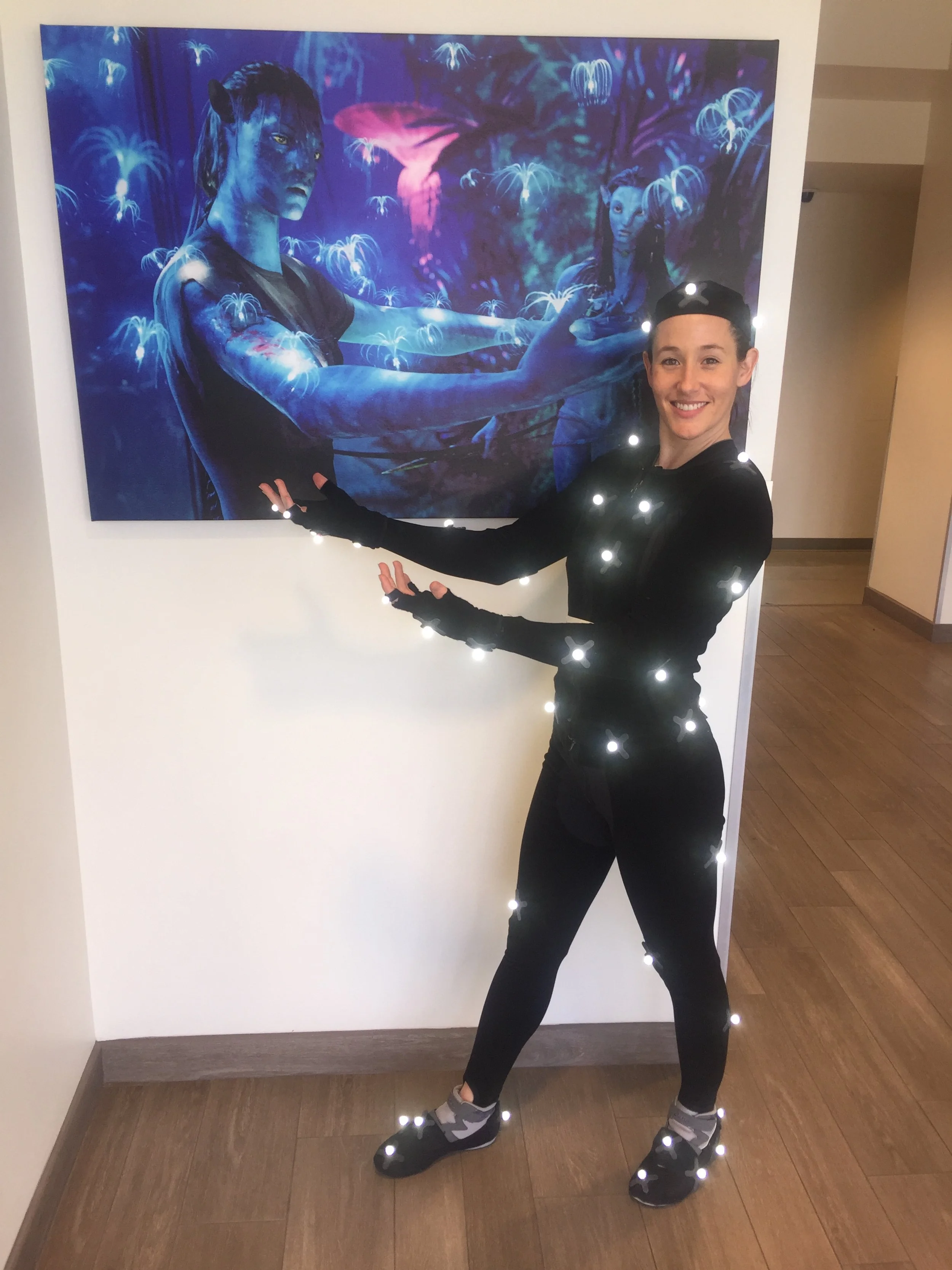 A woman in black athletic clothing and black shoes stands in front of a colorful Avatar artwork, with her arms extended, presenting the artwork. Her clothing is embedded with motion capture markers.