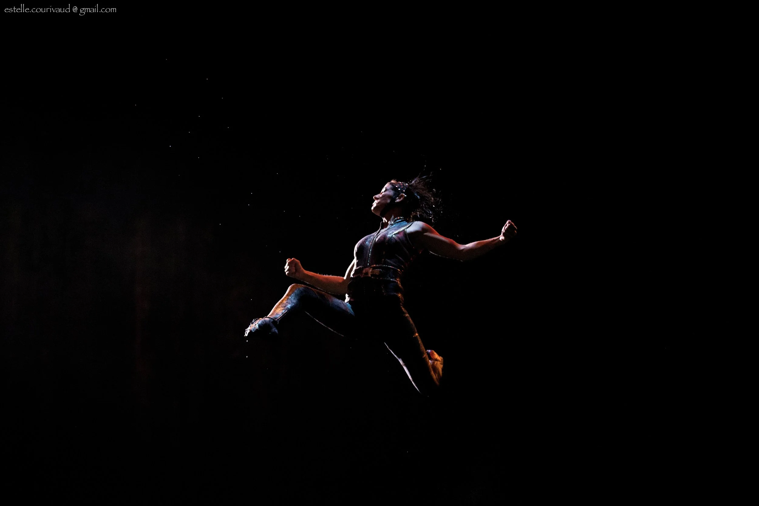 A performer jumping off Russian swing into a dark space, with water droplets around her. She is wearing athletic clothing and has her hair loose.