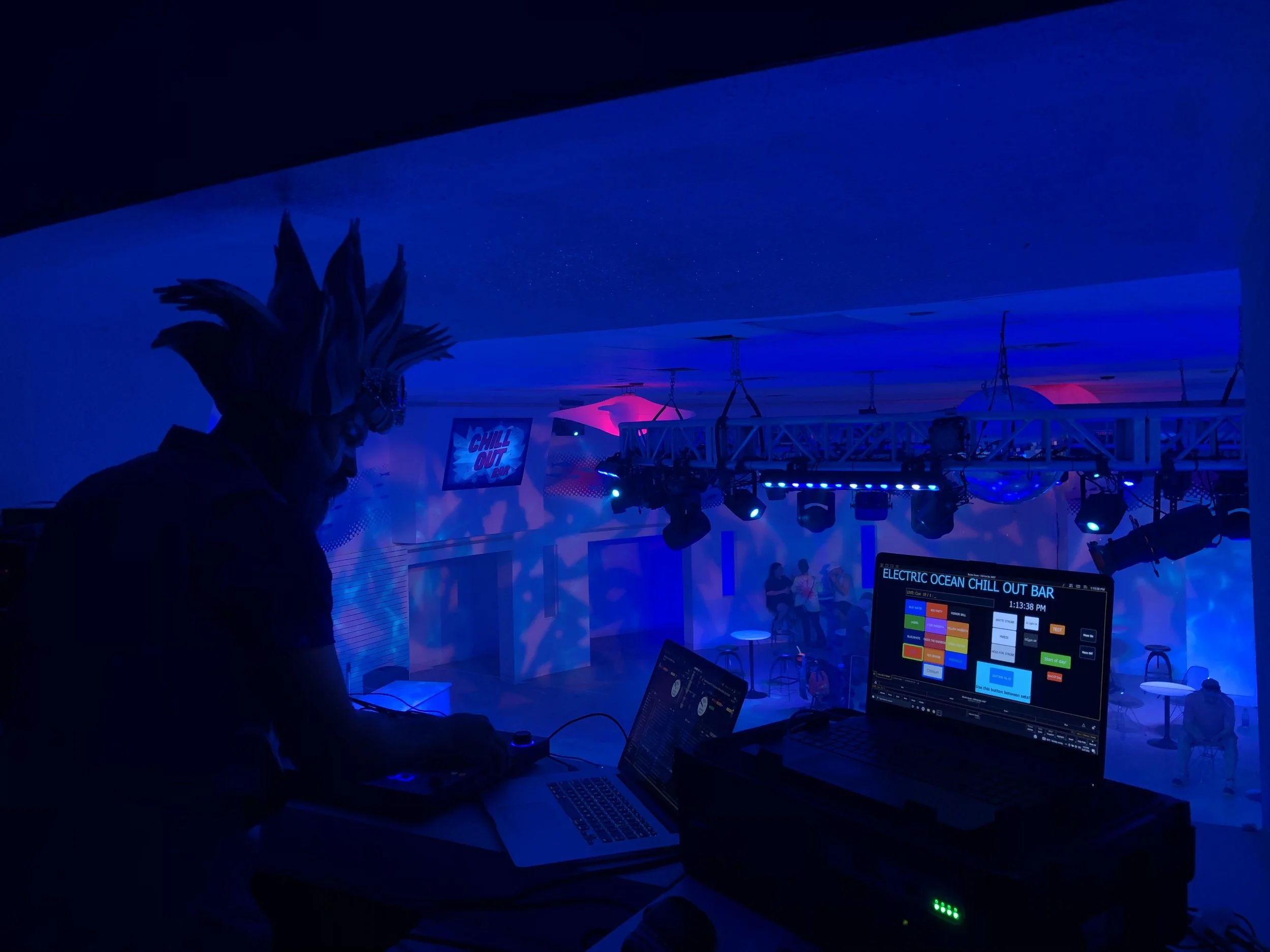 A DJ in a dark room with blue lighting and a costume with spiky hair, working on a laptop and DJ equipment at an electric chill out bar, with a dance floor and people in the background.