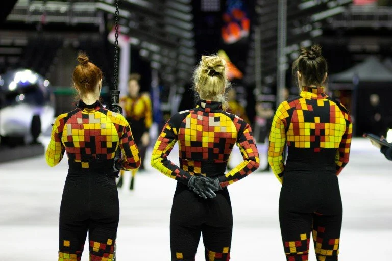 Three women with their backs to the camera, wearing matching yellow, red, and black pixel-pattern costumes in an ice skating arena.