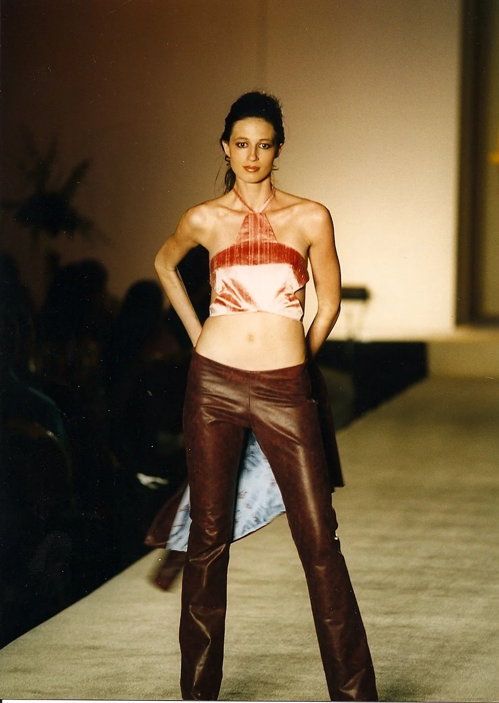 A female model walking on a fashion runway wearing a pink satin halter top and burgundy leather pants.
