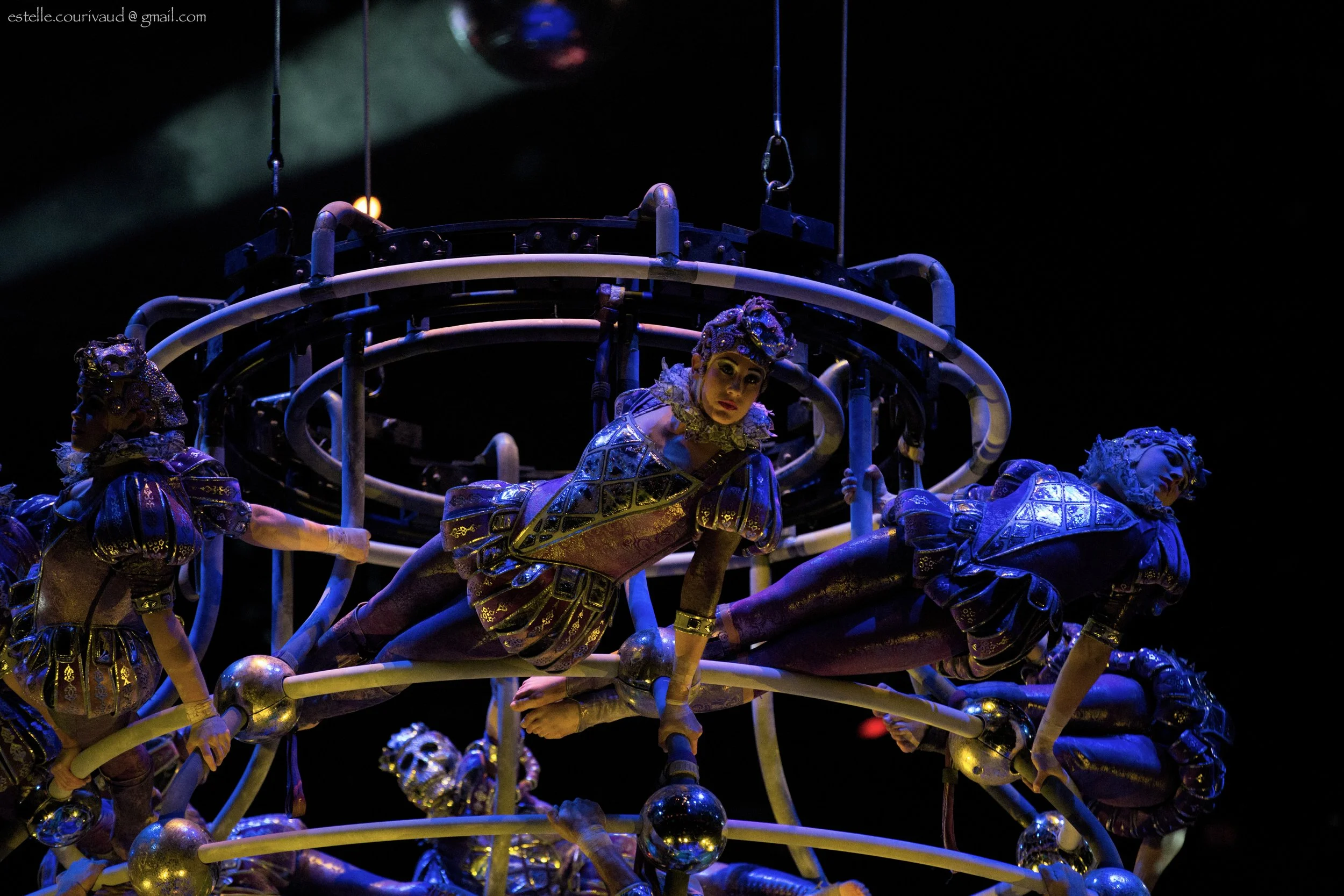 Performer dressed in elaborate costume, lying on a metallic structure, surrounded by dark stage lighting with a spotlight highlighting her face, during a theatrical or dance performance.