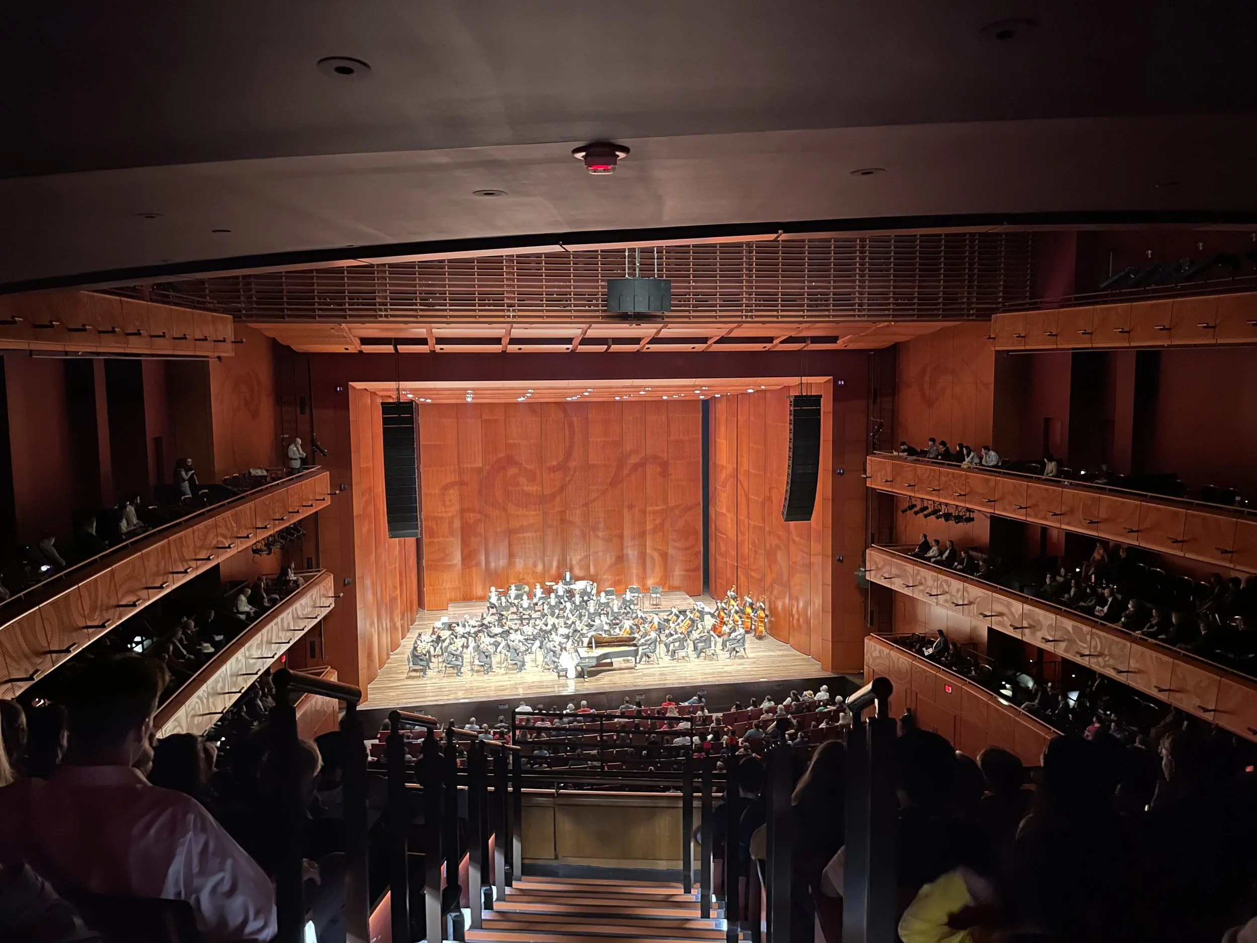 View of an auditorium with a live orchestral performance on stage, with audience seated in the lower section and balconies on both sides.