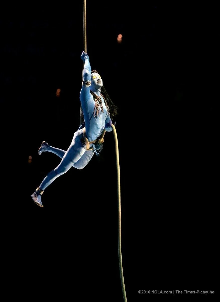 Aerial performer dressed as a character from Avatar, with blue skin and tribal makeup, hanging from a rope during a stage performance in an indoor arena against a dark background.