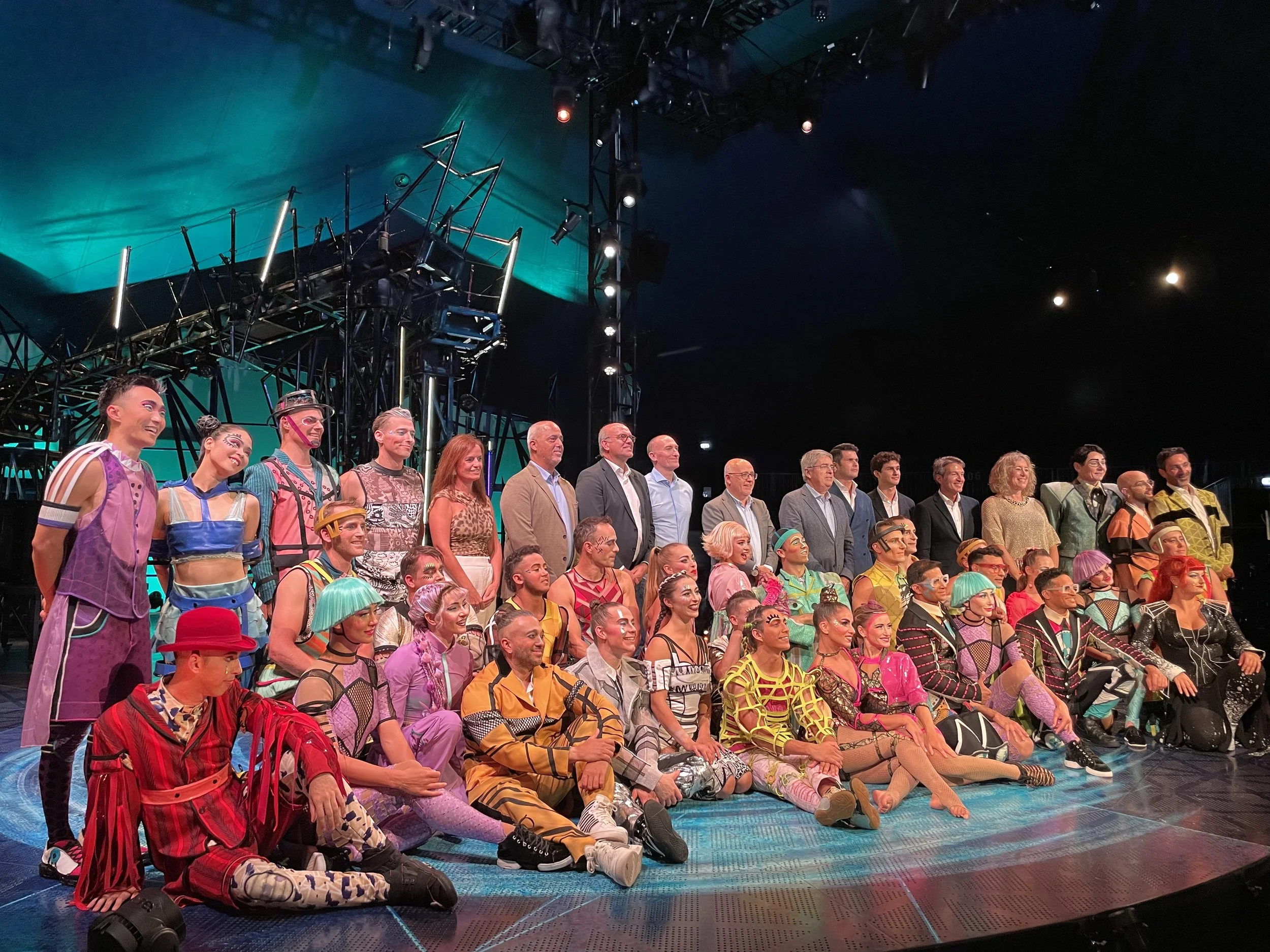 Group photo of performers dressed in colorful costumes and a few in casual suits, posing on a stage after a performance, with stage equipment and lighting visible in the background.