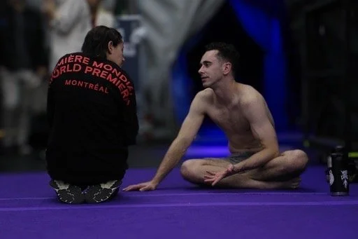 A man sits cross-legged on the floor, talking to a woman who has her back to the camera, wearing a black shirt with red text on the back.