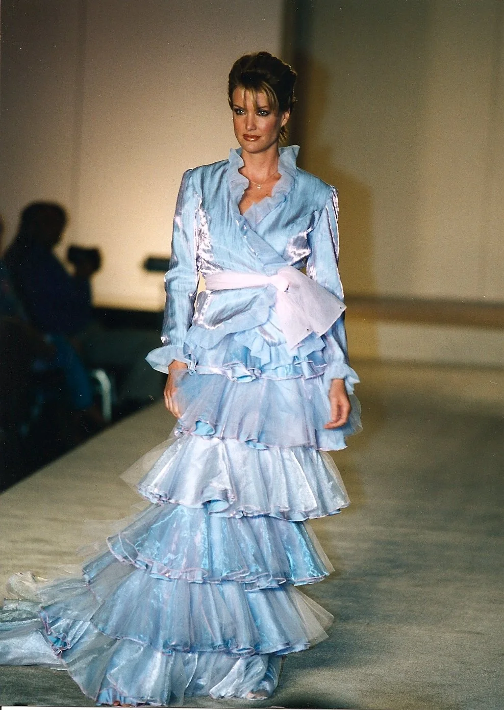 A female model walks on a runway wearing a shiny, layered, light blue gown with ruffles, tied at the waist with a wide ribbon.