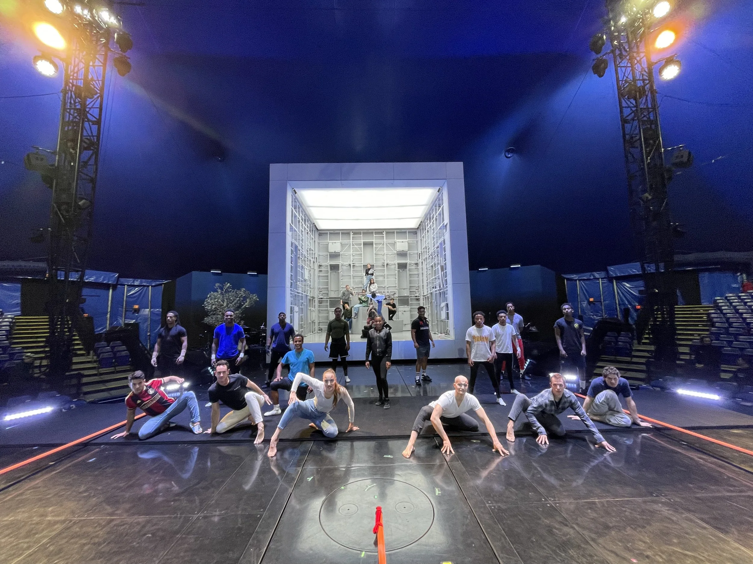 Group of performers on stage practicing for a performance, with some kneeling or crouching in front, others standing in the background, stage lighting and a large central cube structure behind them.