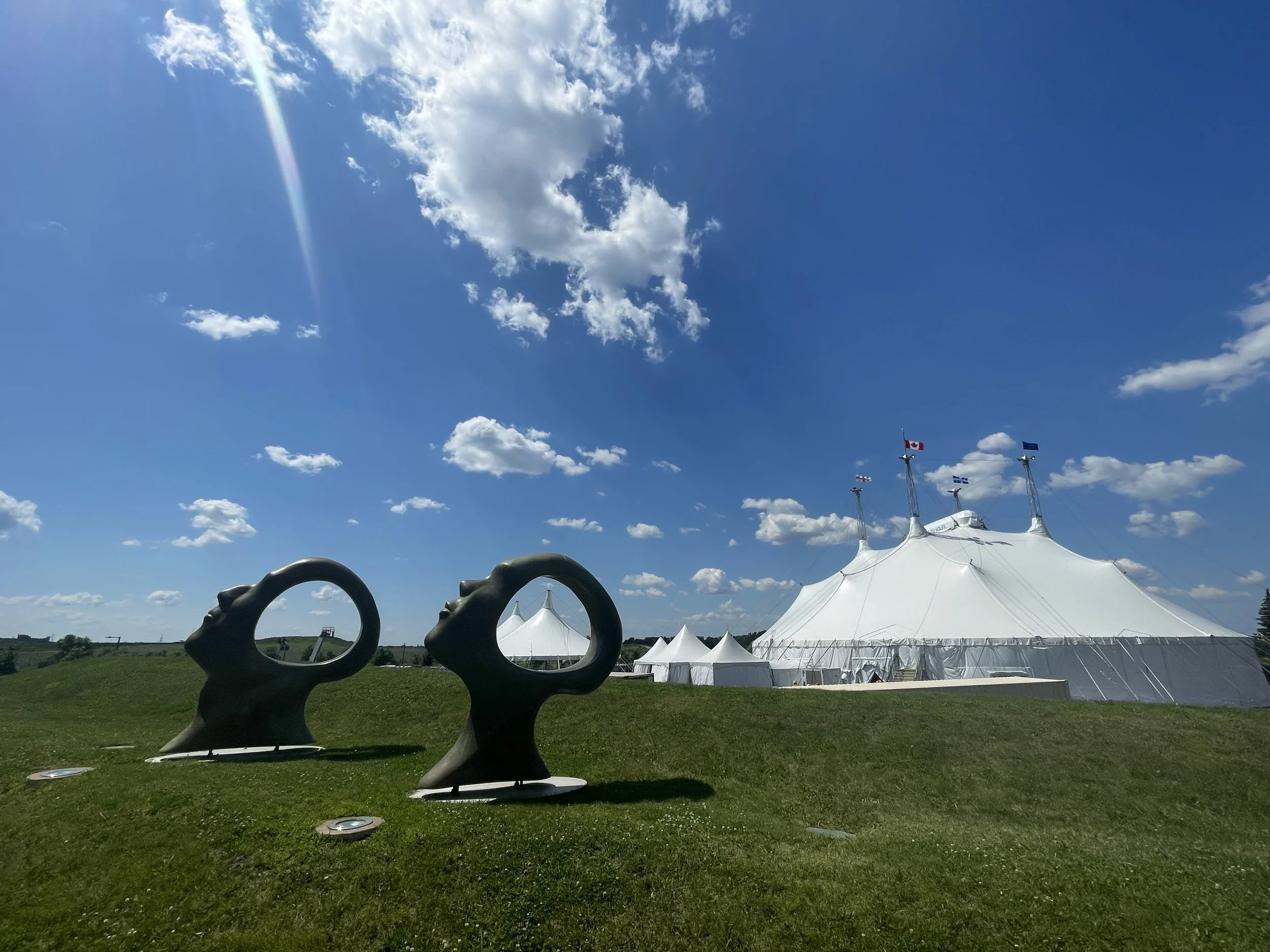 A large white circus tent with flagpoles on top, surrounded by smaller tents, on a grassy hill under a bright blue sky with scattered clouds and a lens flare at Cirque du Soleil International Headquarters.