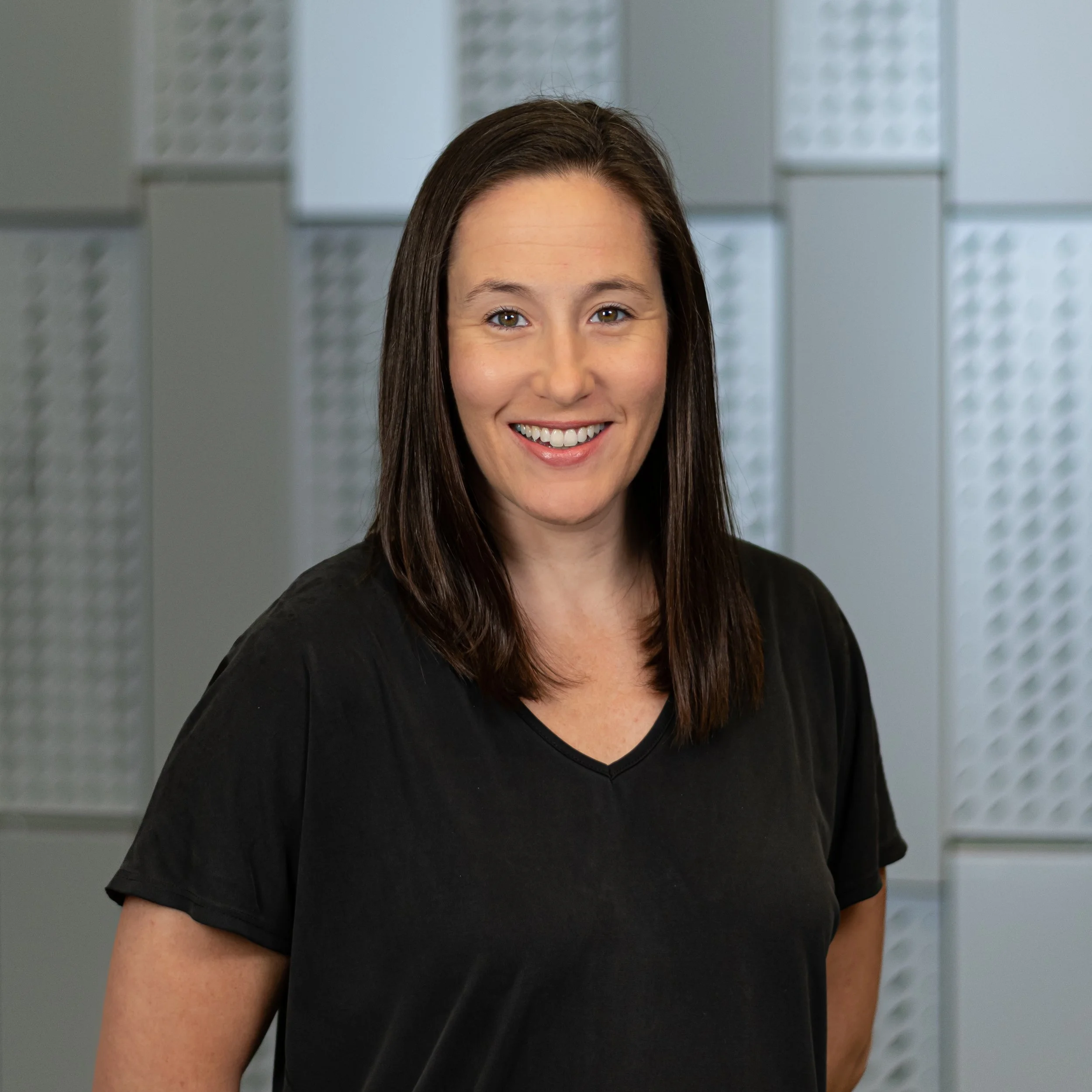 A woman with shoulder-length brown hair, smiling, wearing a black t-shirt, standing in front of a textured, patterned wall.