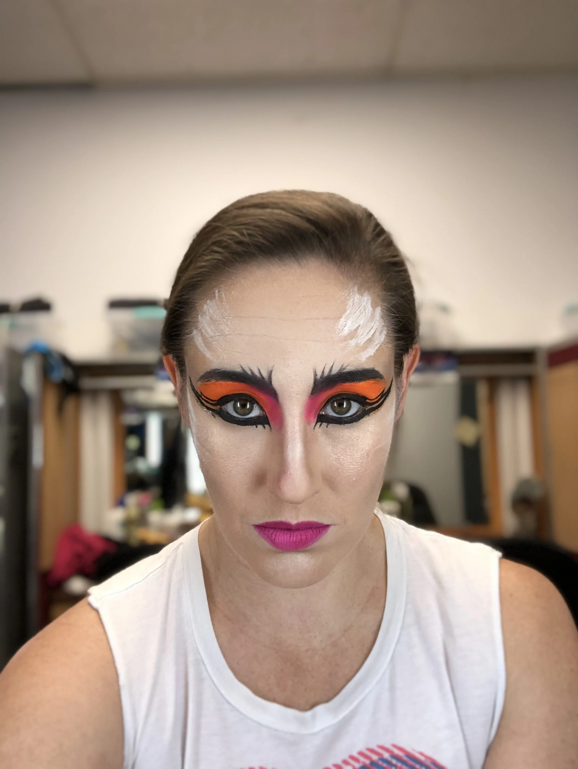 Female with bold and colorful dramatic makeup, including orange, pink, and black eyeshadow, winged eyeliner, pink lipstick, and white accents on forehead, wearing a white sleeveless top, in a room with shelves and blurred background.