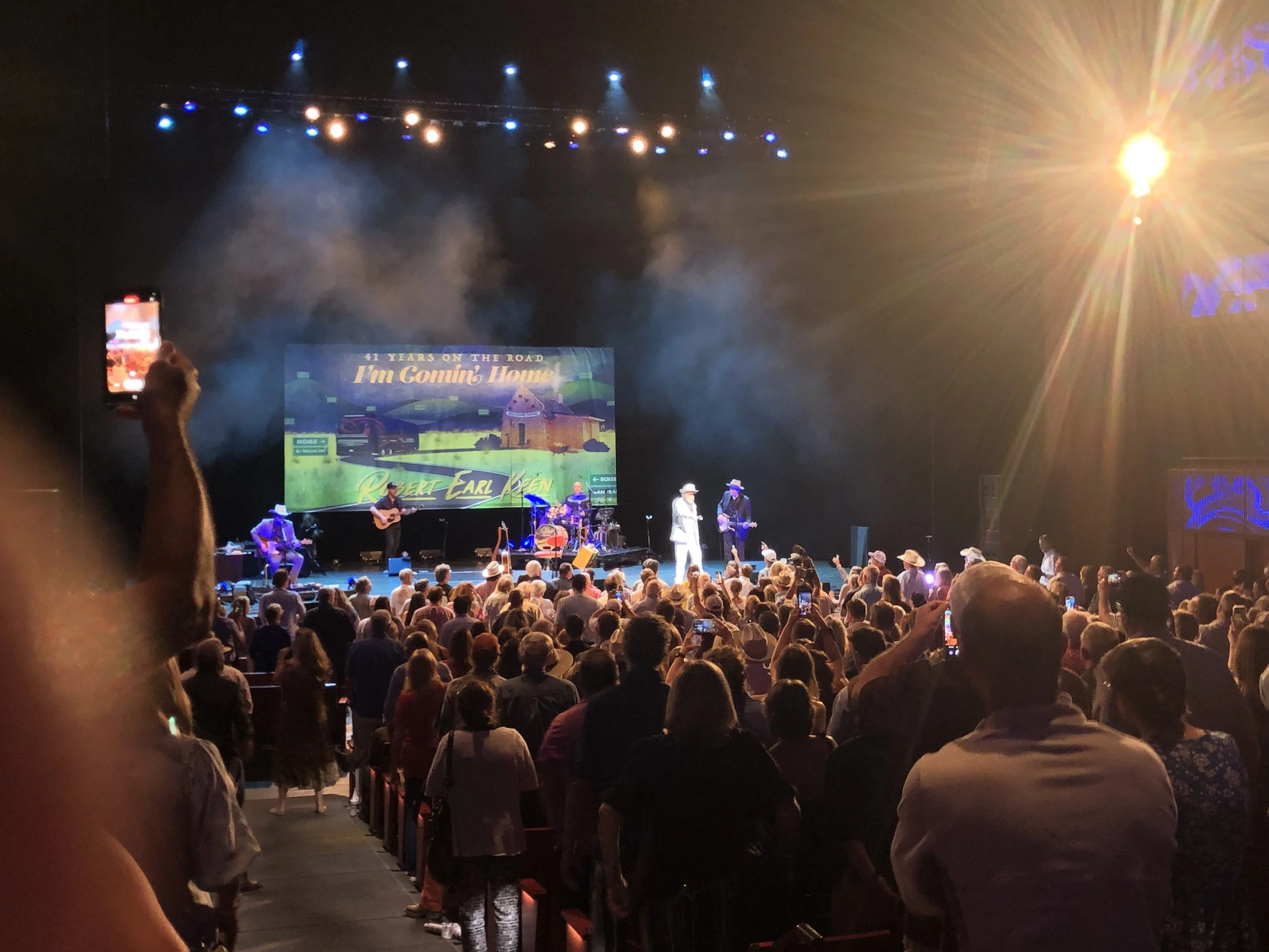 Concert stage with performers, audience watching, and large screen in the background.