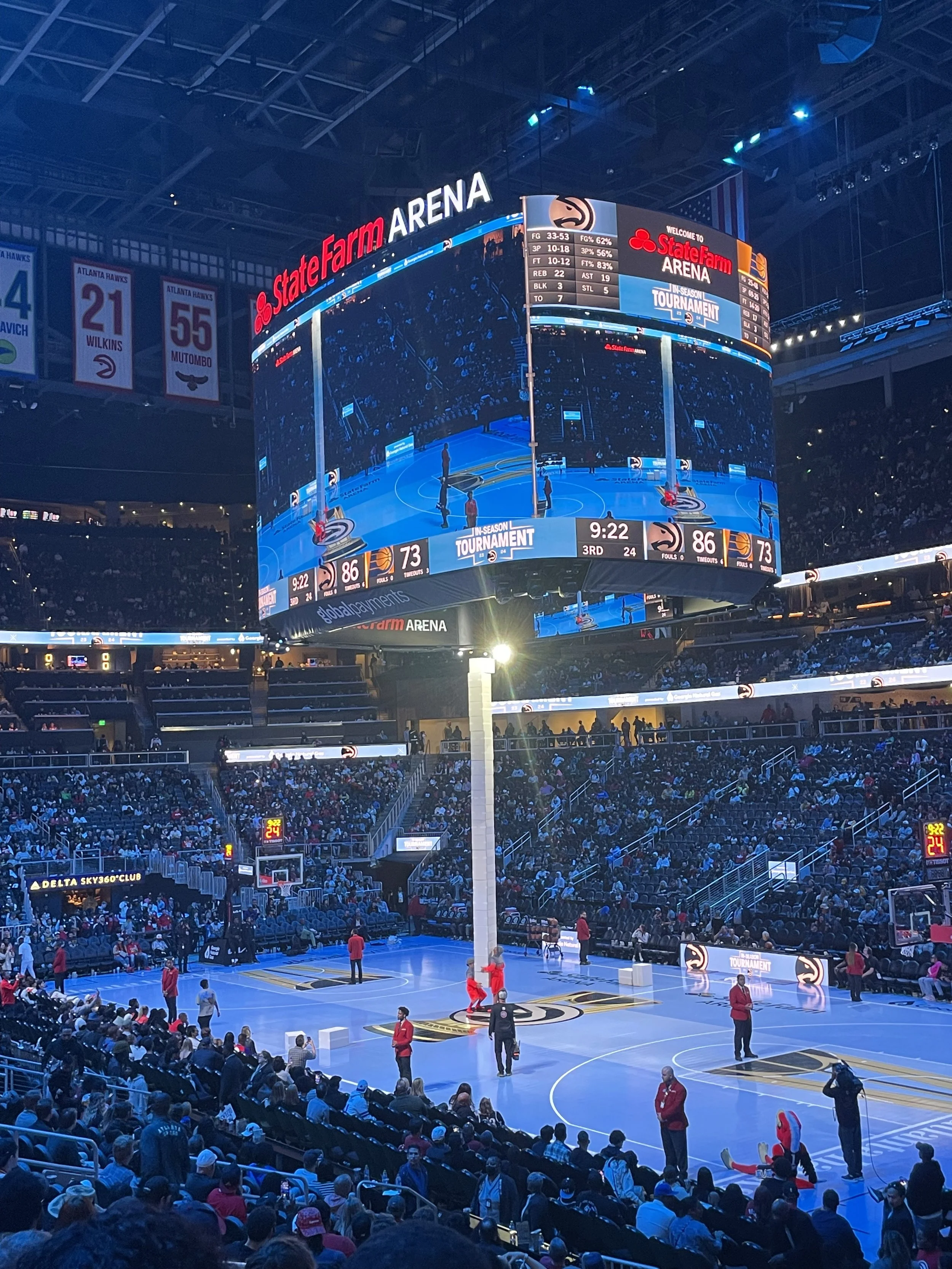 A basketball arena filled with spectators, a court with a half time show performance, and a large digital scoreboard displaying game scores and time.
