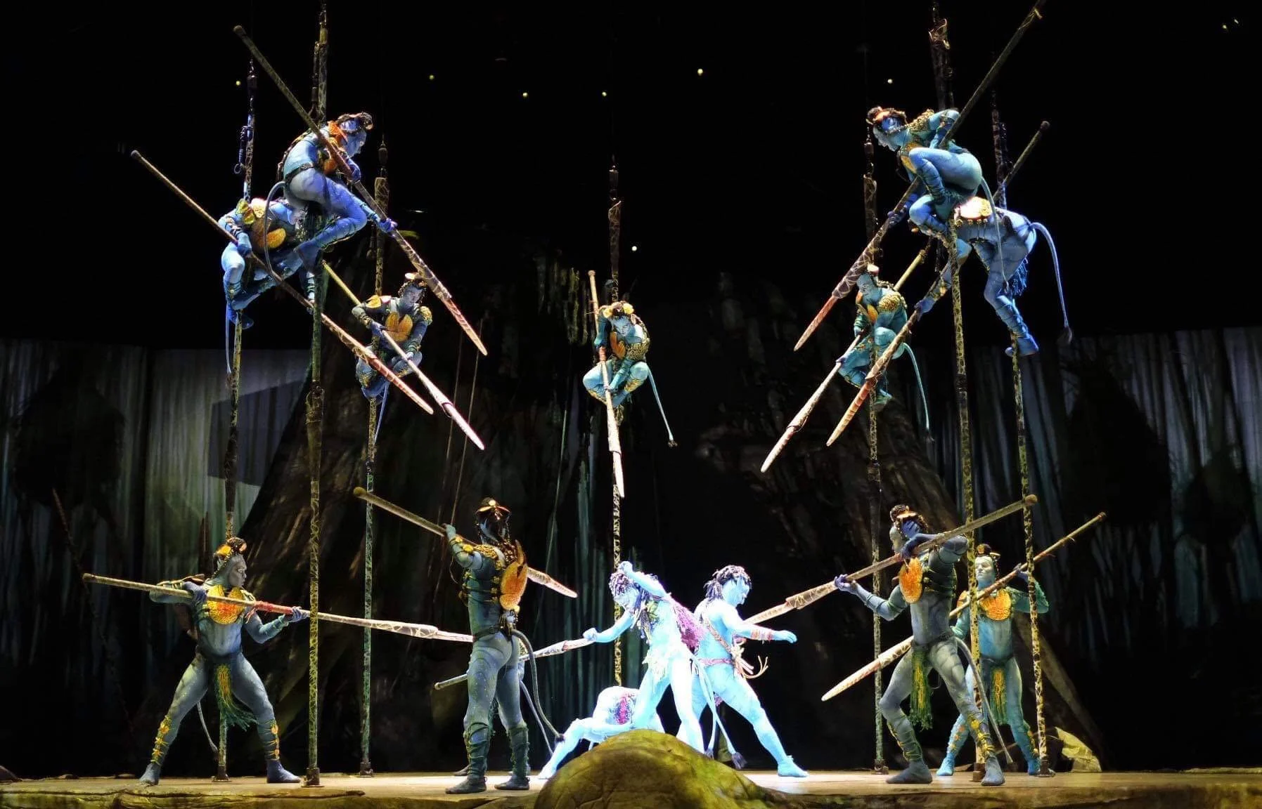 Performers dressed as characters from Avatar on a stage with some performers on the poles and some on the ground, mid-performance, under stage lighting.