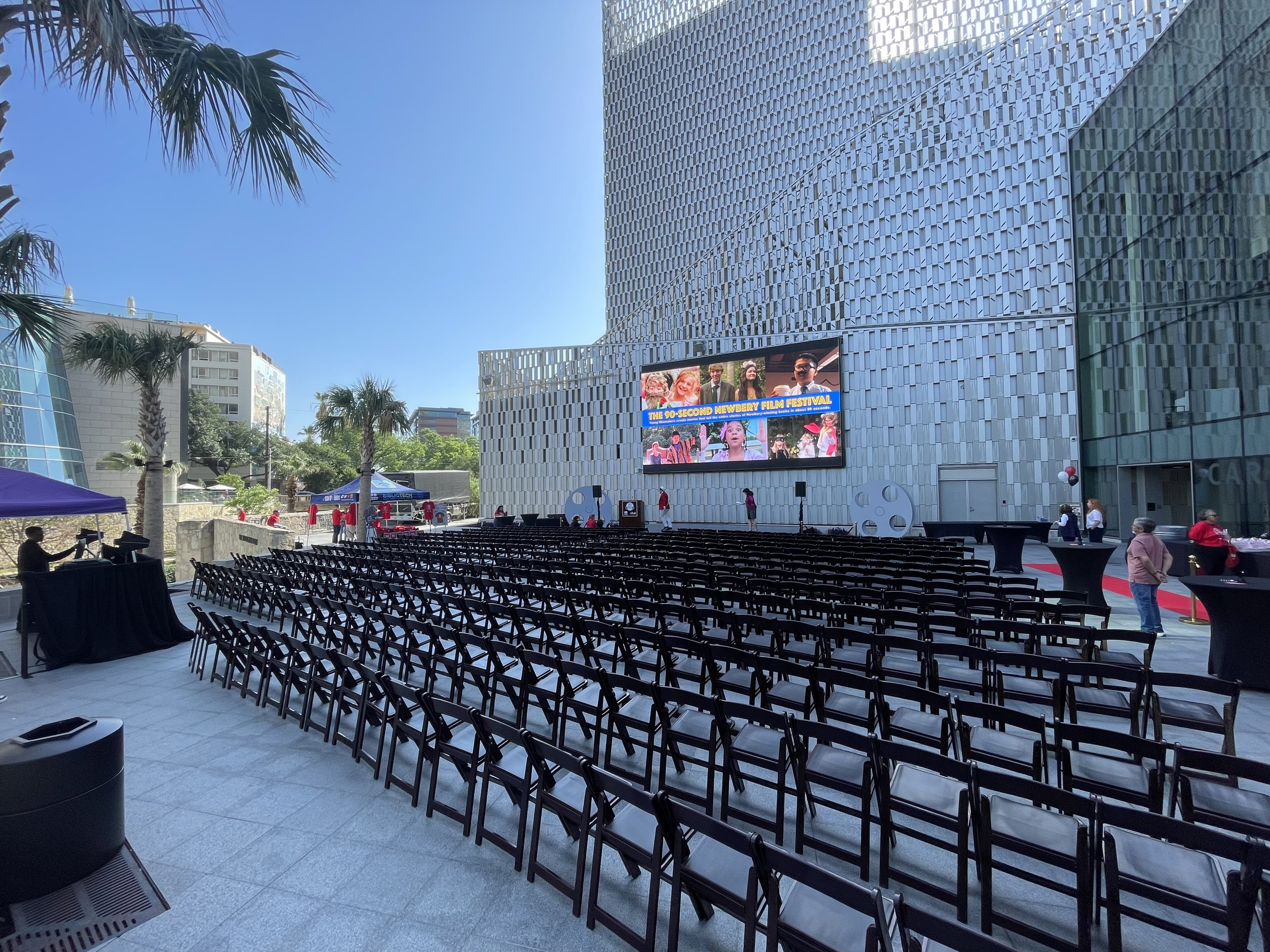 An outdoor event setup with rows of chairs facing a large digital screen on a modern building. There are a few people setting up and preparing, with some palm trees in the background and a clear blue sky.