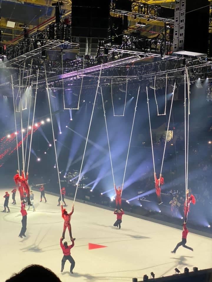 Circus performers in red costumes holding long chains, performing on an ice rink with stage lighting and scaffolding overhead.