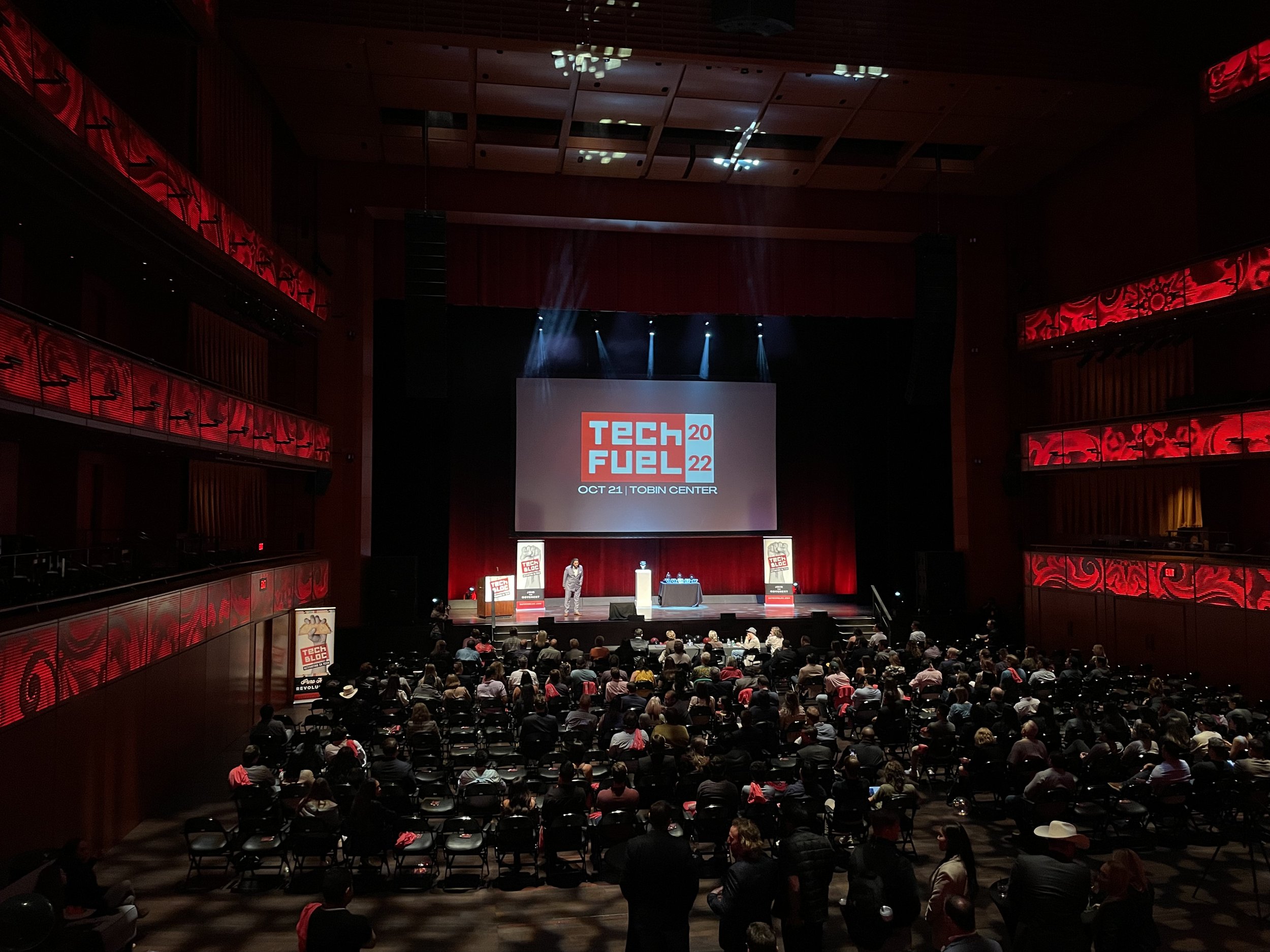 A large conference hall filled with seated audience watching a presentation on a stage. The stage has a large screen displaying 'TECH FUEL 2022' with the date October 21 at Tobin Center. There are speakers, banners, and red decorative lighting around