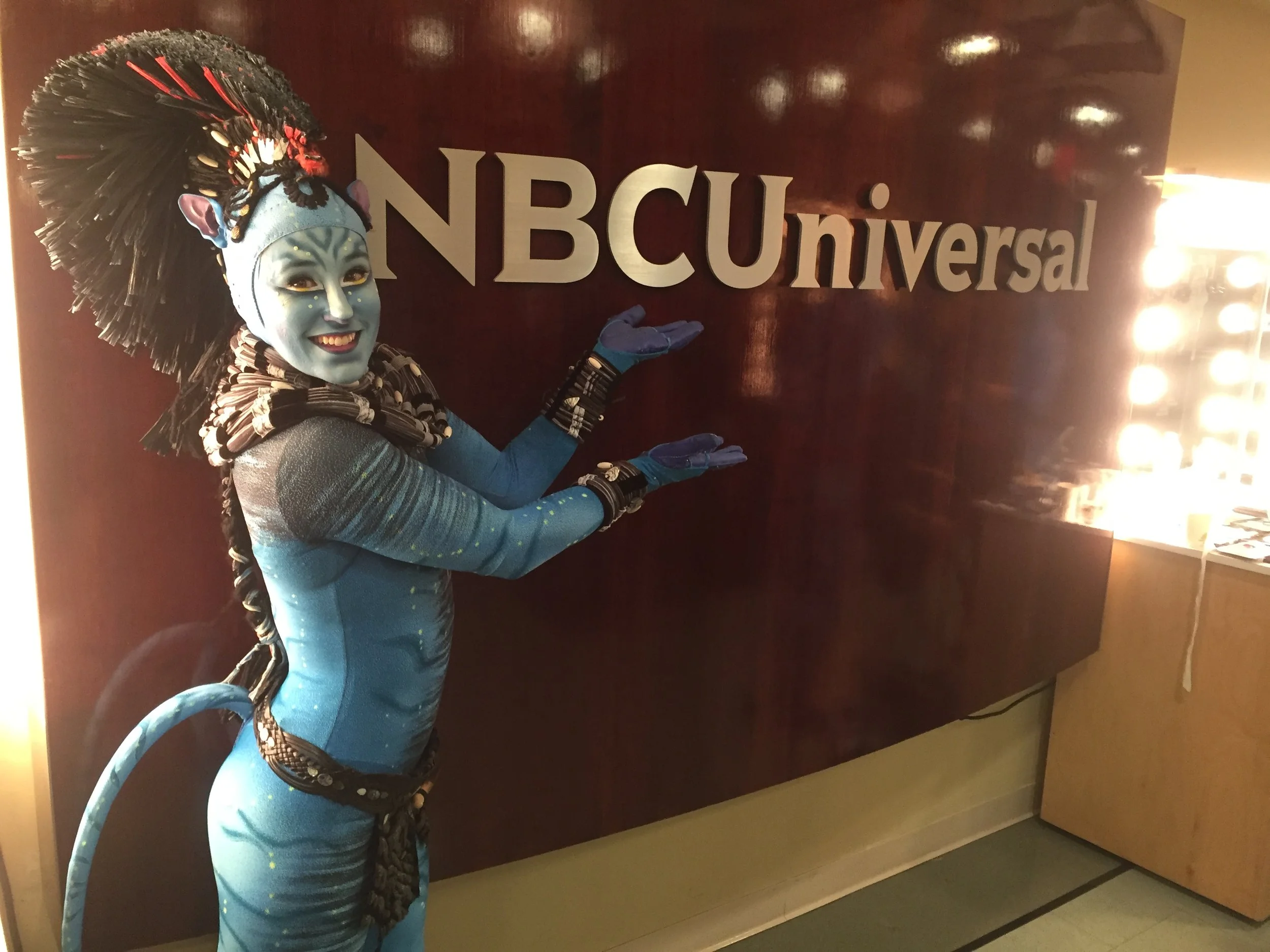 A person dressed as a Navi from Avatar at NBCUniversal sign, wearing blue body paint, a headpiece, and costume accessories.