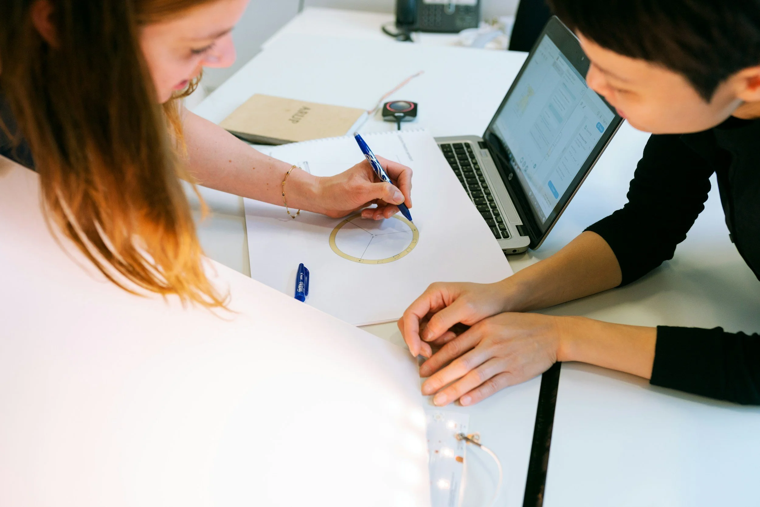 Two people are working together at a white table, reviewing a printed diagram with a color wheel and a ruler, alongside an open laptop showing charts, notebooks, and office supplies in the background.