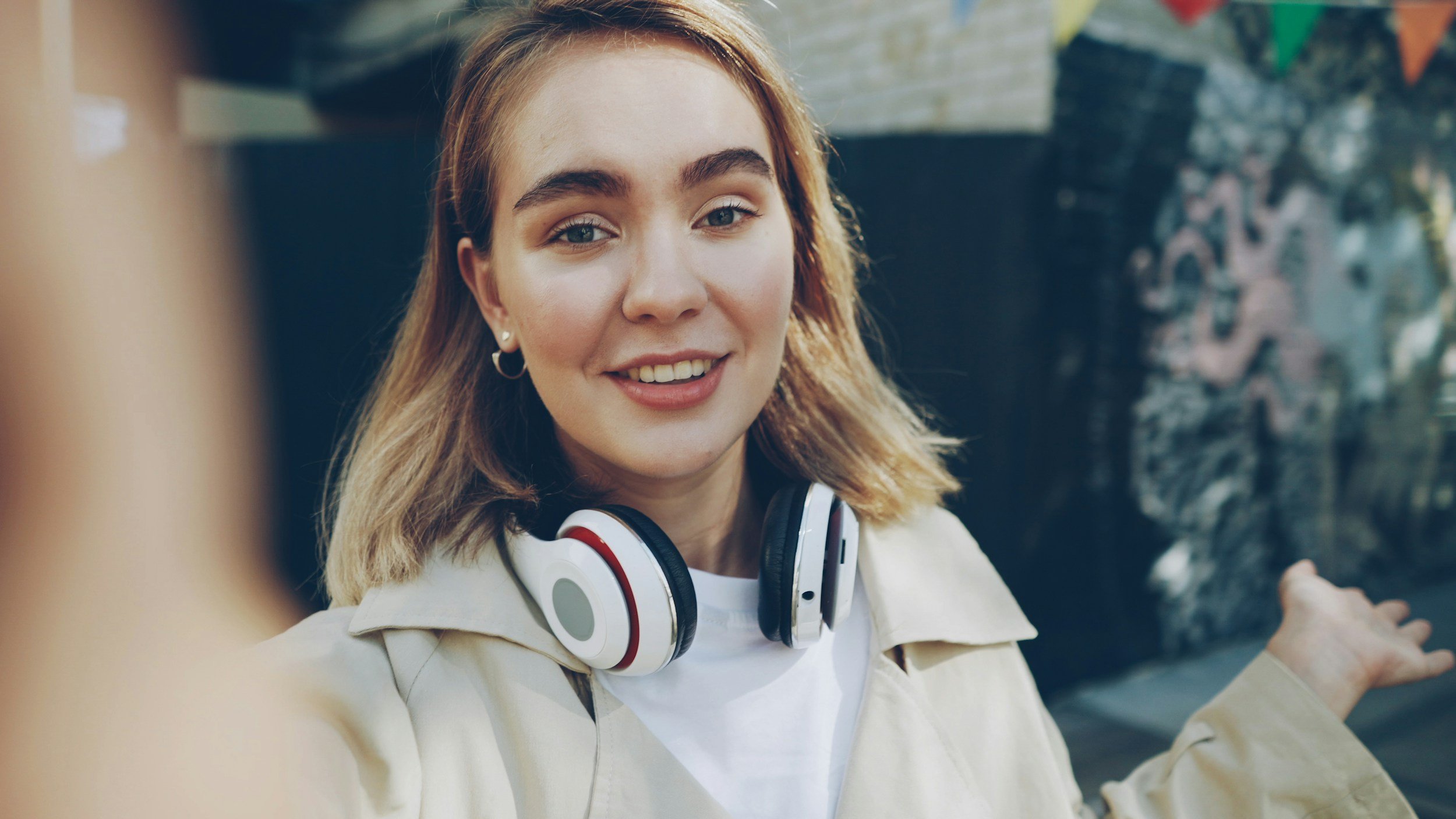 A young woman with shoulder-length blond hair smiling at the camera, wearing a beige jacket, white t-shirt, and large white and red headphones around her neck, standing outdoors near water with trees and buildings in the background.