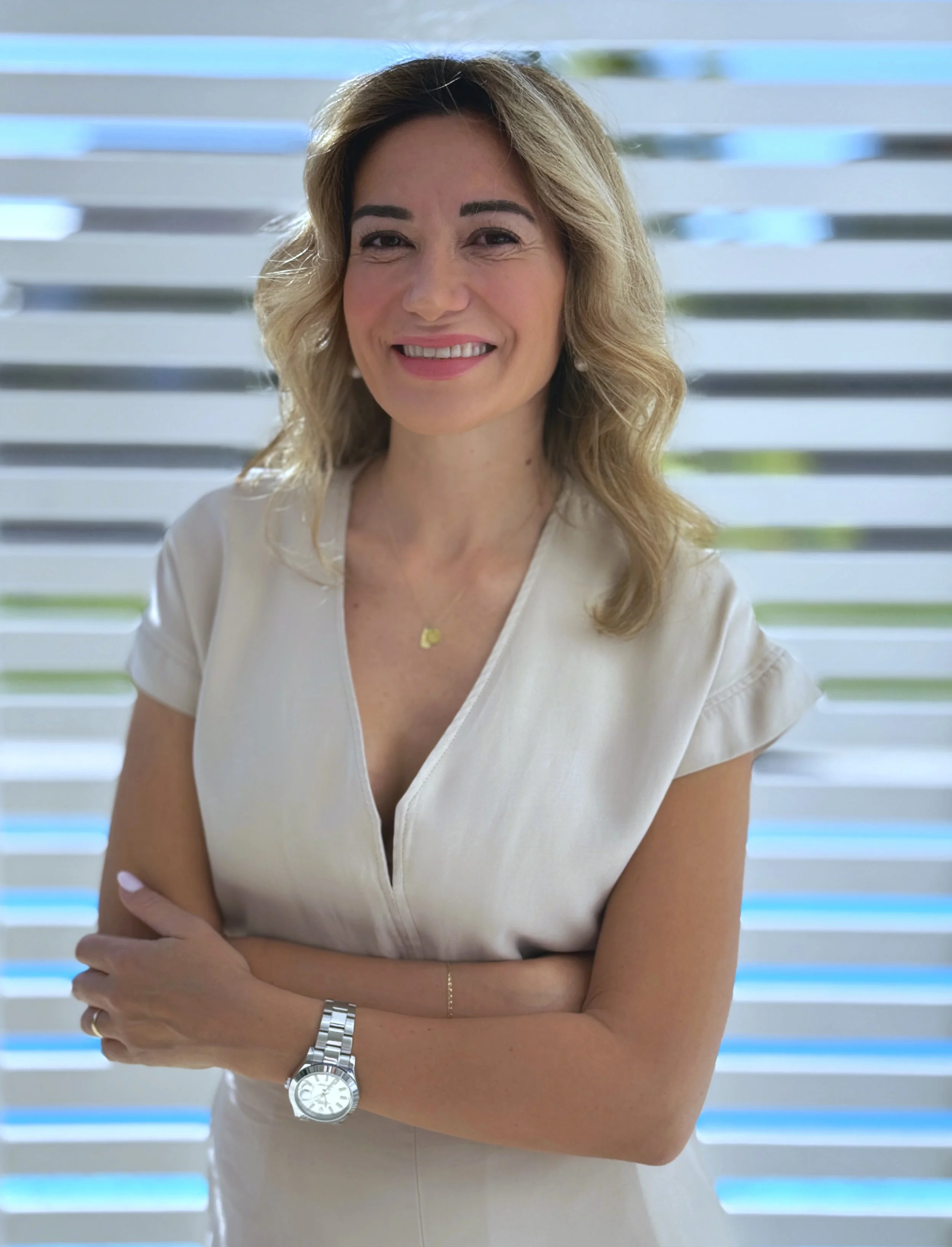 Eine Frau mit blonden, gewellten Haaren, die vor einem modernen, lamellenartigen Hintergrund steht, lächelnd und in einem weißen Kleid.