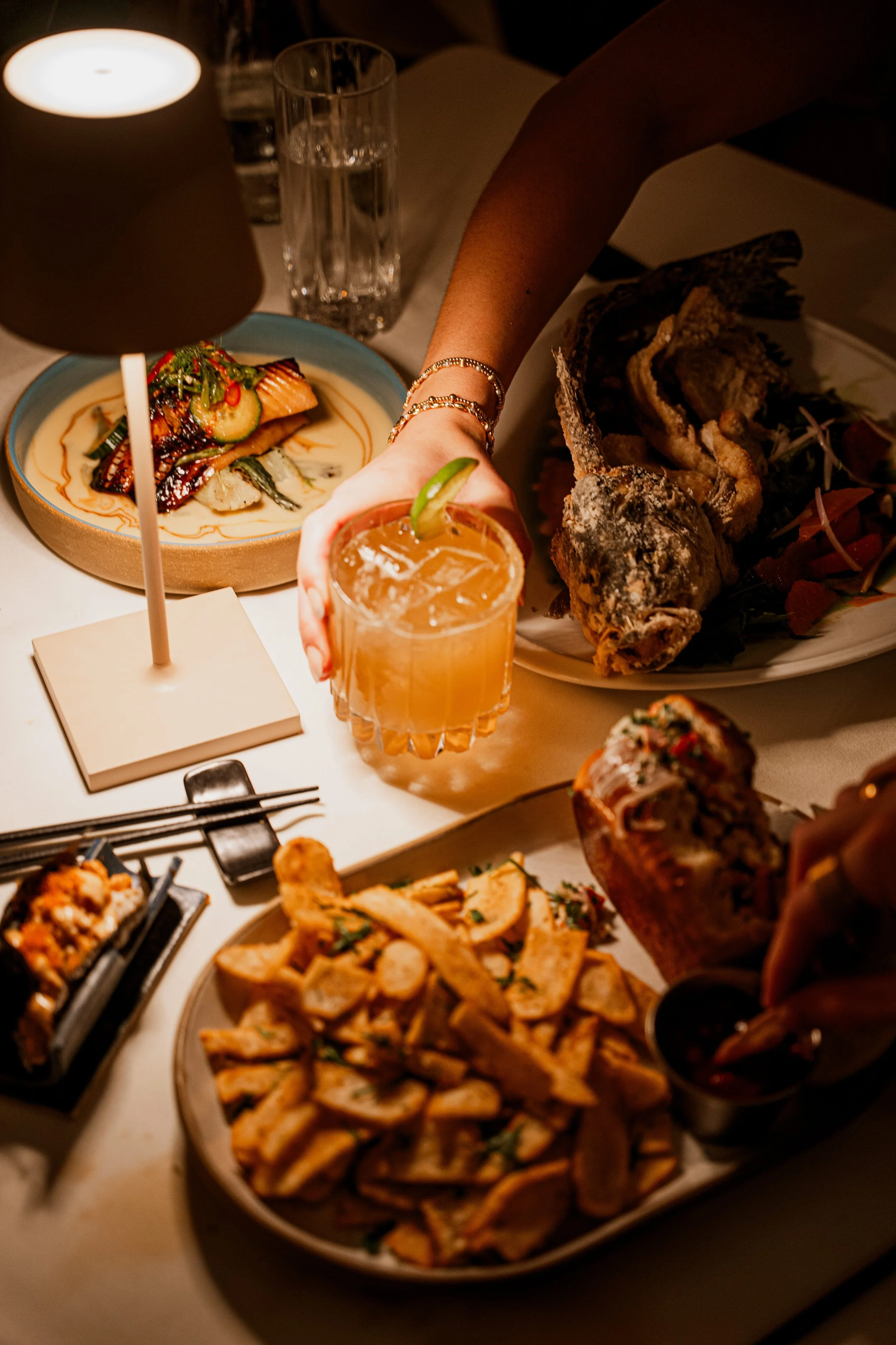 Party table with various dishes and drinks, including a large fried fish, a plate of seasoned chips, a cocktail with lime, and other plated foods.