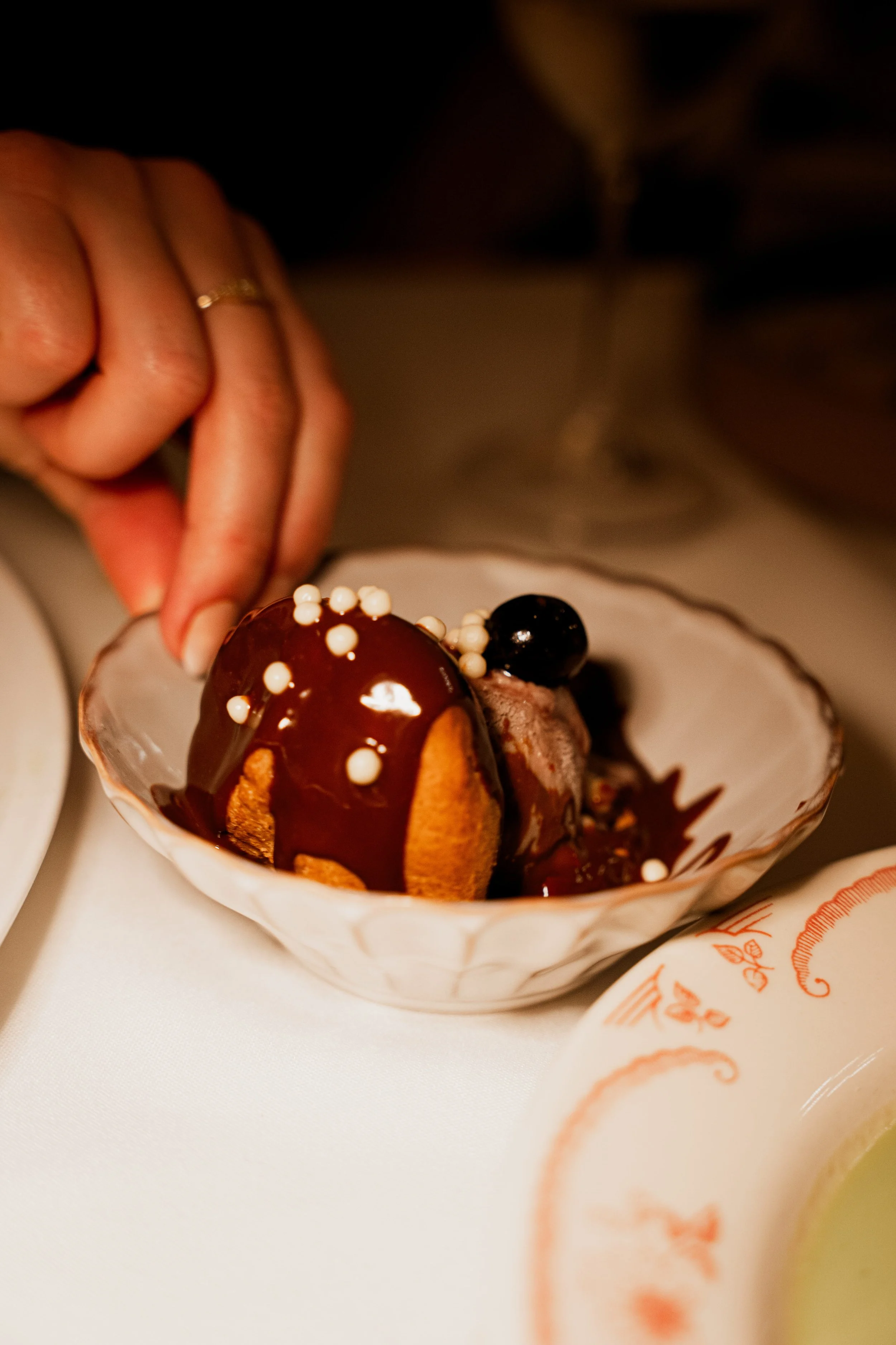 A person's hand with a wedding ring holding a spoon over a bowl of ice cream topped with chocolate sauce, white sprinkles, and a blueberry.