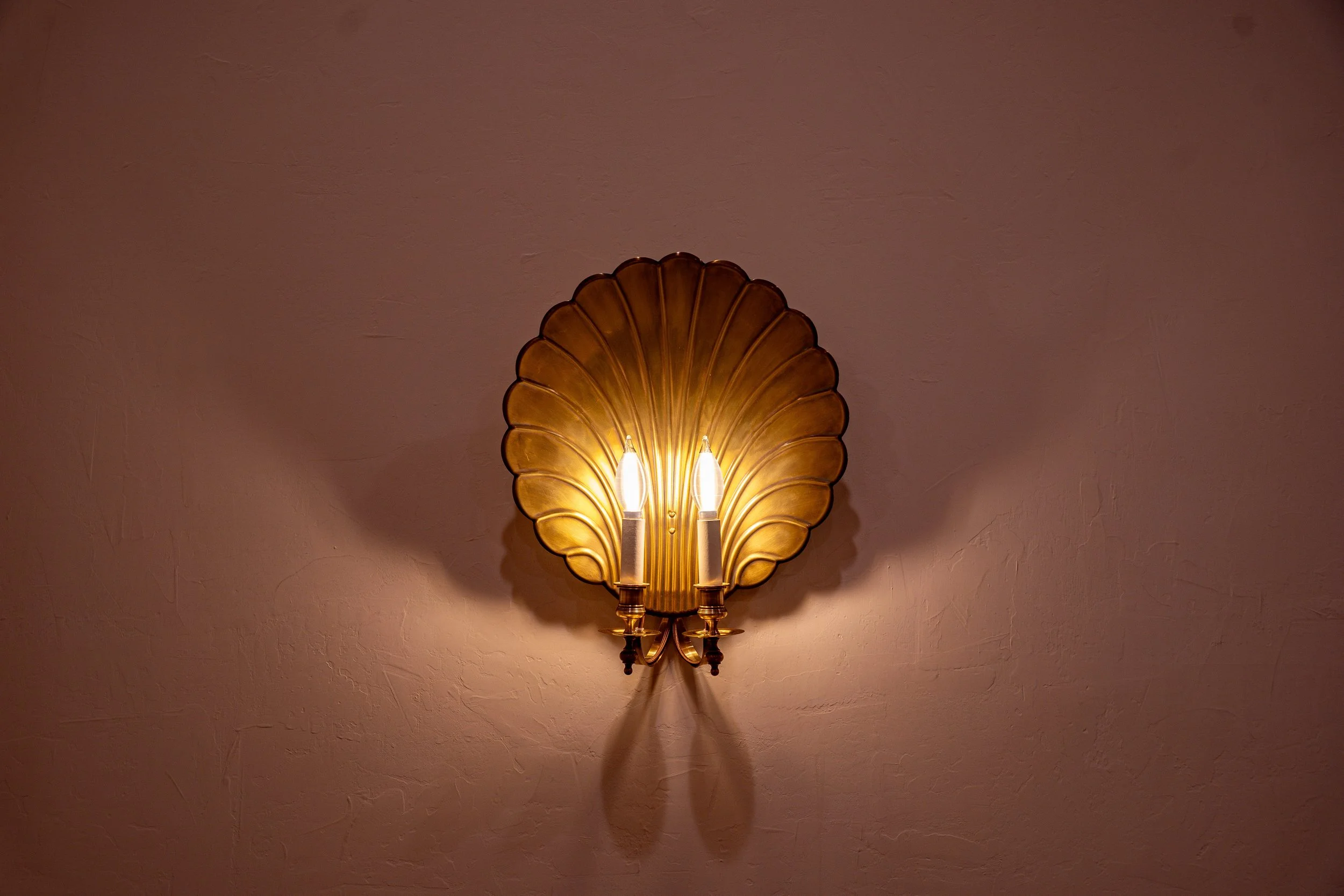A vintage wall sconce with two lights, mounted on a textured beige wall. The sconce has a decorative gold-colored metal backplate shaped like a leaf or shell, with two candle-like light bulbs.