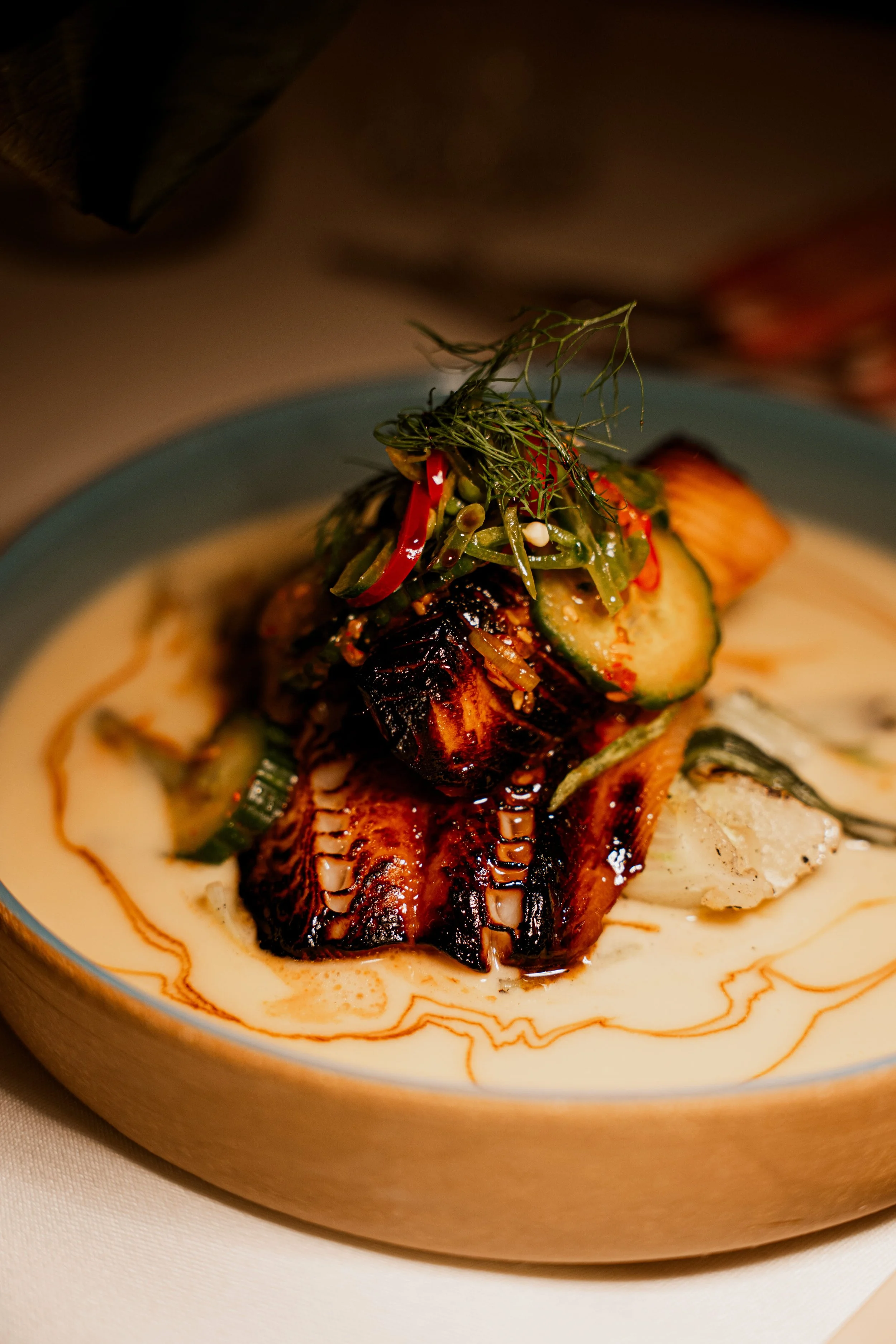 A plated dish featuring grilled octopus with a creamy sauce, topped with grilled zucchini, red bell peppers, and fresh herbs.