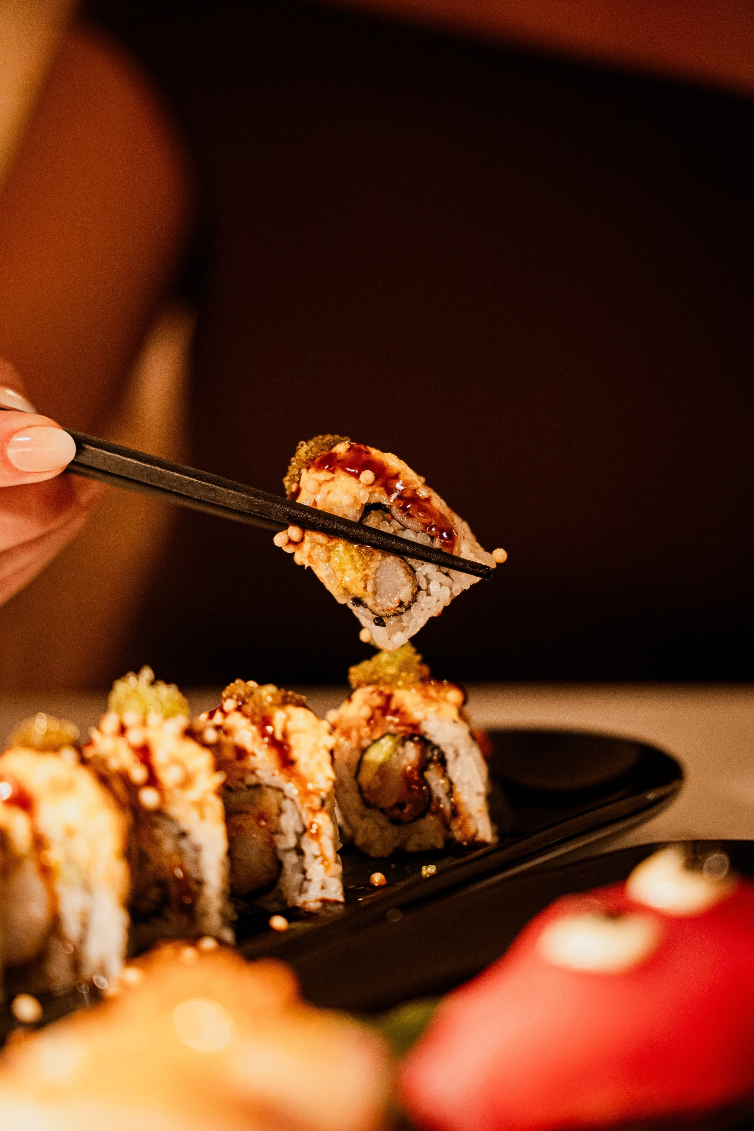 A person holding chopsticks picking up a piece of sushi from a black plate with other sushi rolls, with a blurred red object in the foreground.