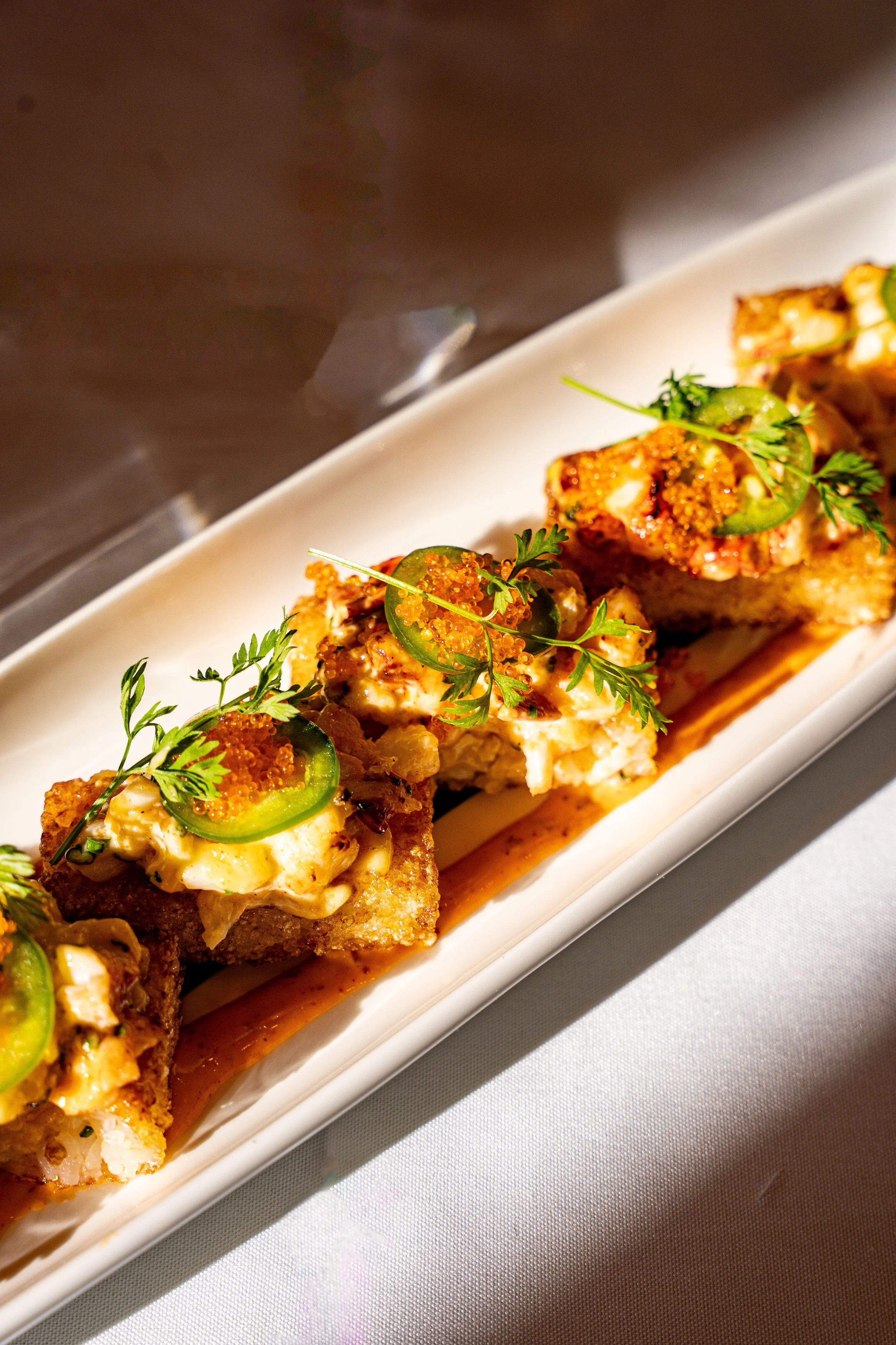 Close-up of sushi rolls garnished with herbs, sliced jalapeños, and orange fish roe, served on a white rectangular plate.