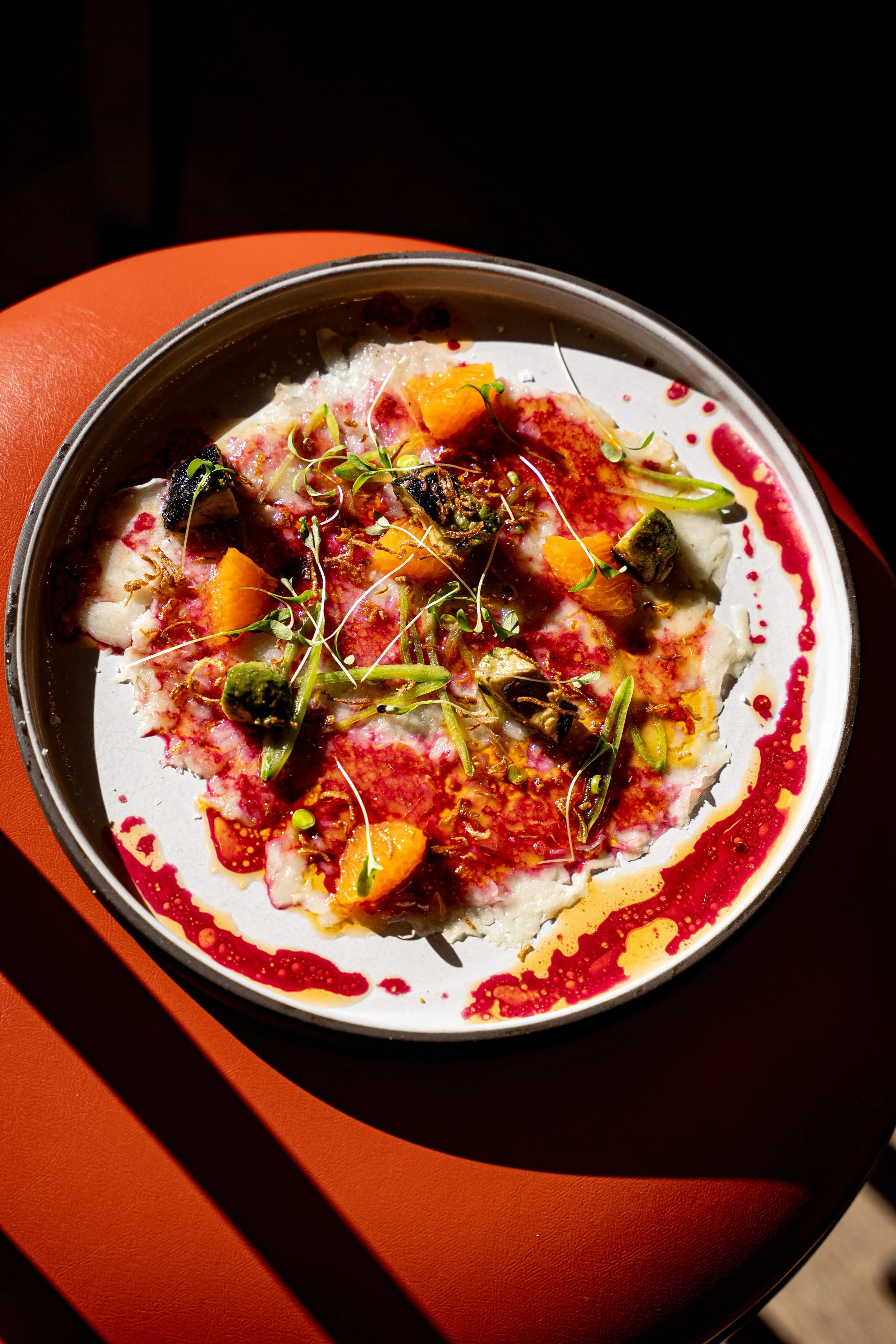 A plated dish with fish, garnished with microgreens and herbs, topped with orange pieces, drizzled with red sauce and oil, on a round dish with background: an orange table.