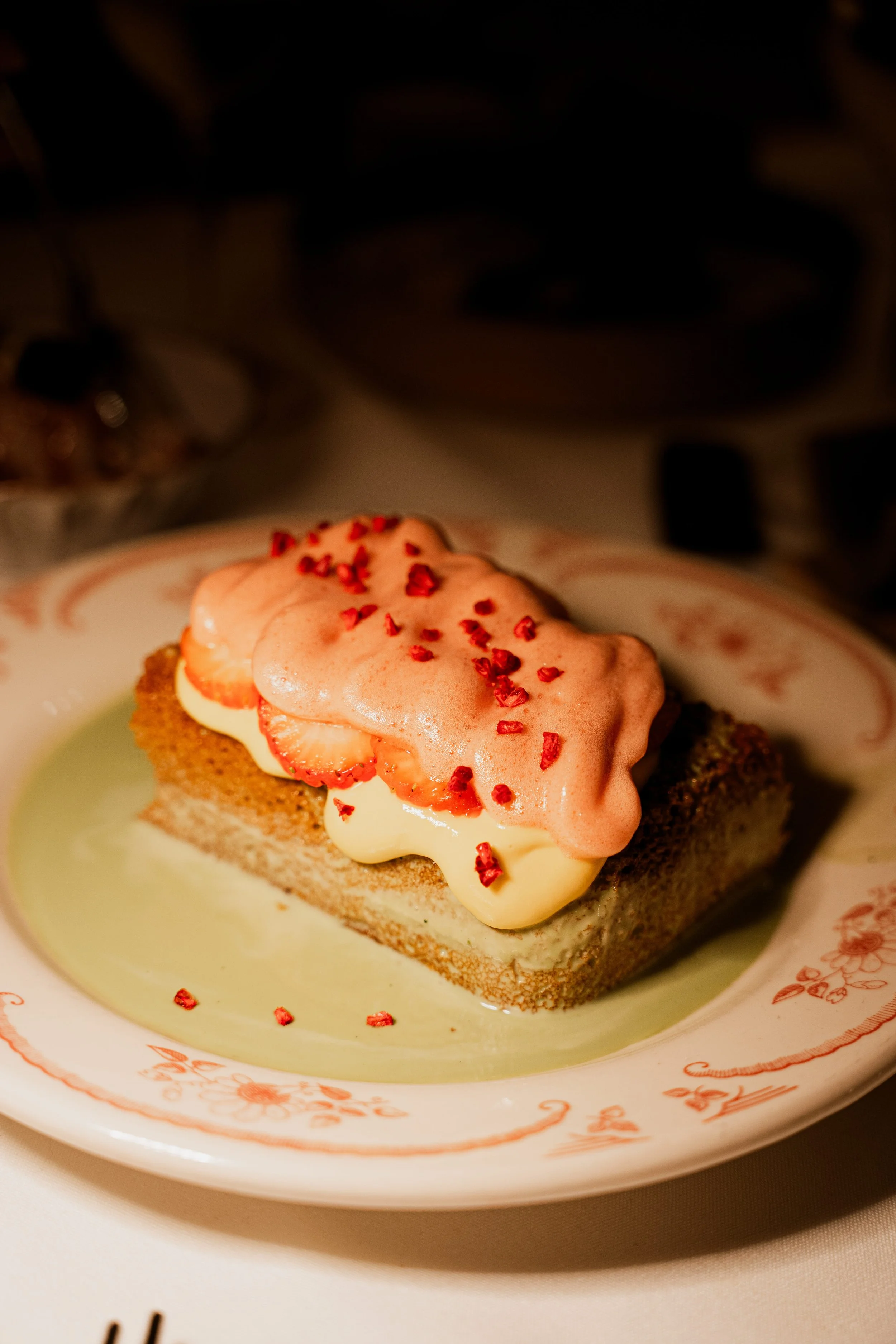 A slice of cake on a decorative plate topped with strawberries, pink whipped cream, and red sprinkles.
