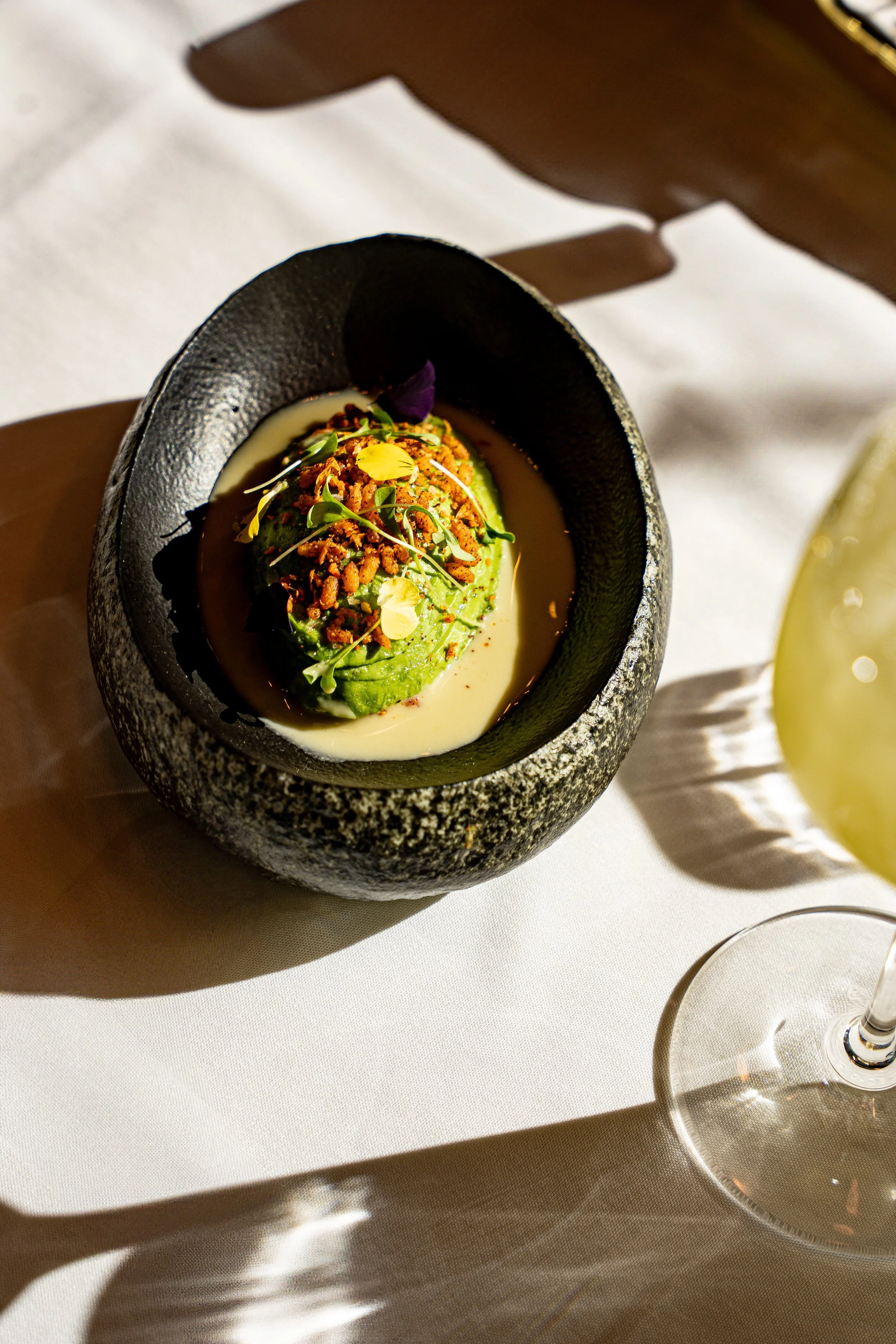 A fancy dish served in a black bowl, featuring small green avocado with toppings like crumbled brown bits and microgreens, placed on a white tablecloth with a glass of white wine nearby.