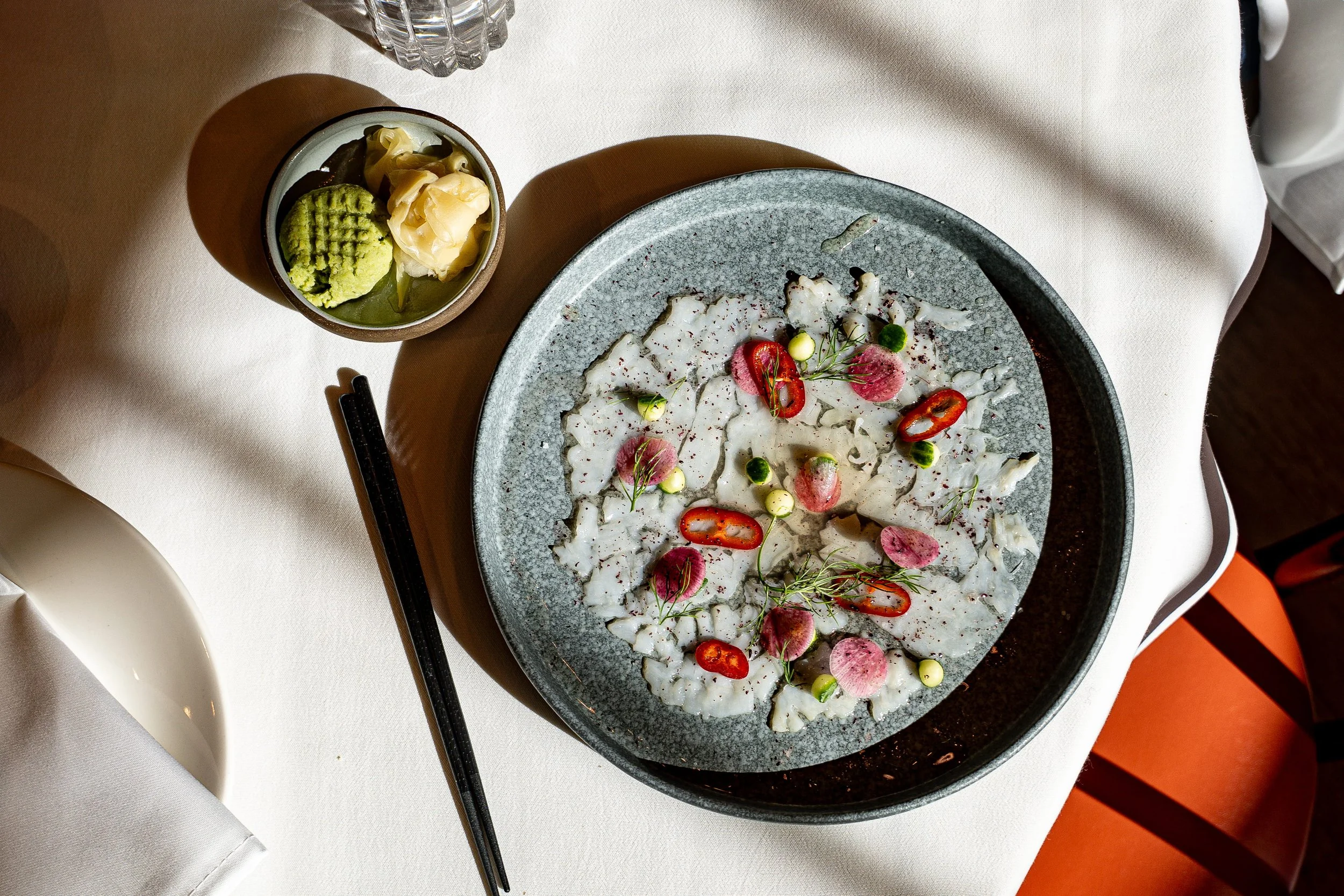 A plate of sashimi garnished with sliced red chili peppers, pink pickled ginger, green herbs, and small green and white vegetables, accompanied by a small bowl of wasabi, pickled ginger, and soy sauce, on a white table with chopsticks and a glass of 